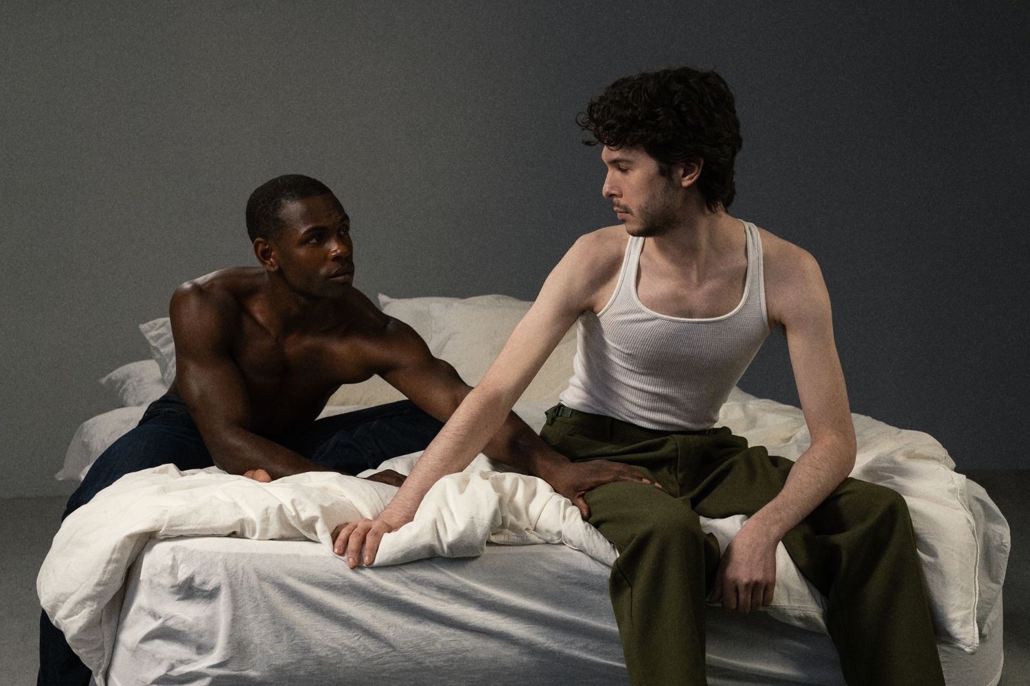Two people sit on a bed in a dramatic scene, one shirtless man and one in a white tank top and green pants.