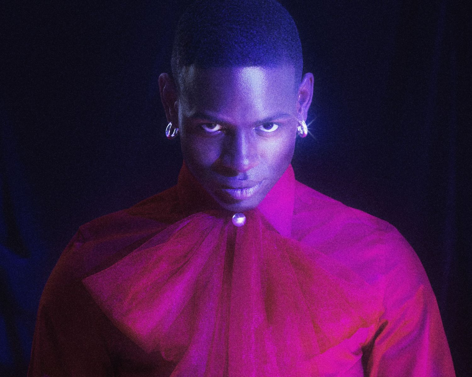 Young man in red sheer bow top bathed in pink and purple neon light in dark setting.