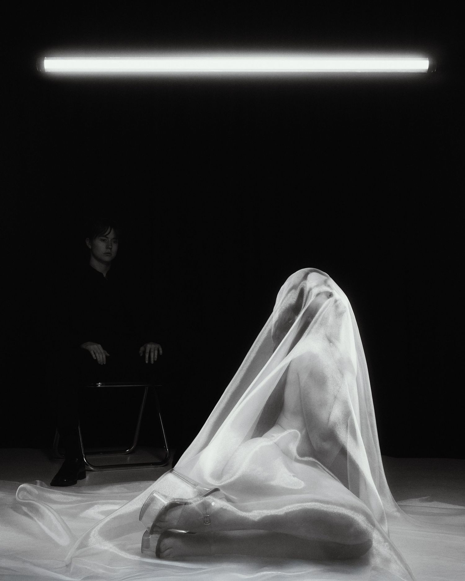 Black and white photo of a person sitting beside a figure draped in sheer fabric under a neon light.