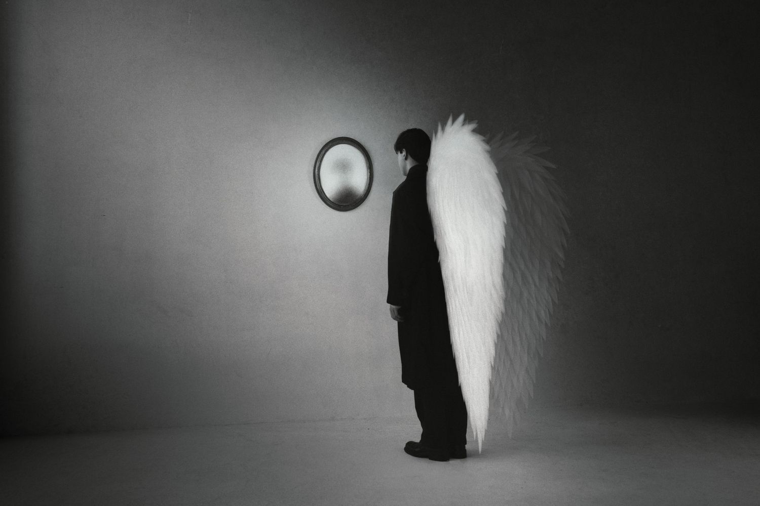 Person in black suit with large white angel wing stands before a round mirror in monochrome.