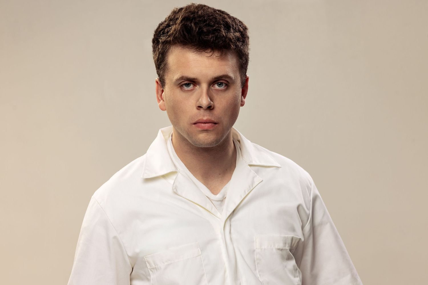 Young man in white button-up shirt posing against a light gray background with a serious expression.