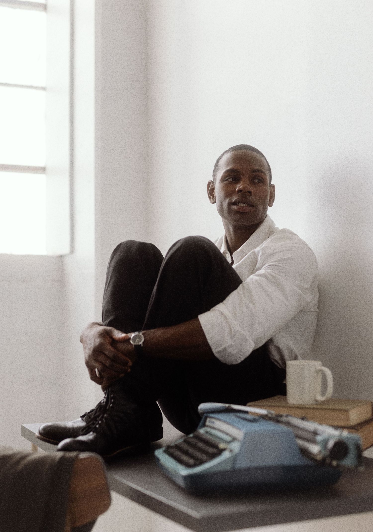 A man in a white shirt sits against a wall beside a blue typewriter, books, and a mug.
