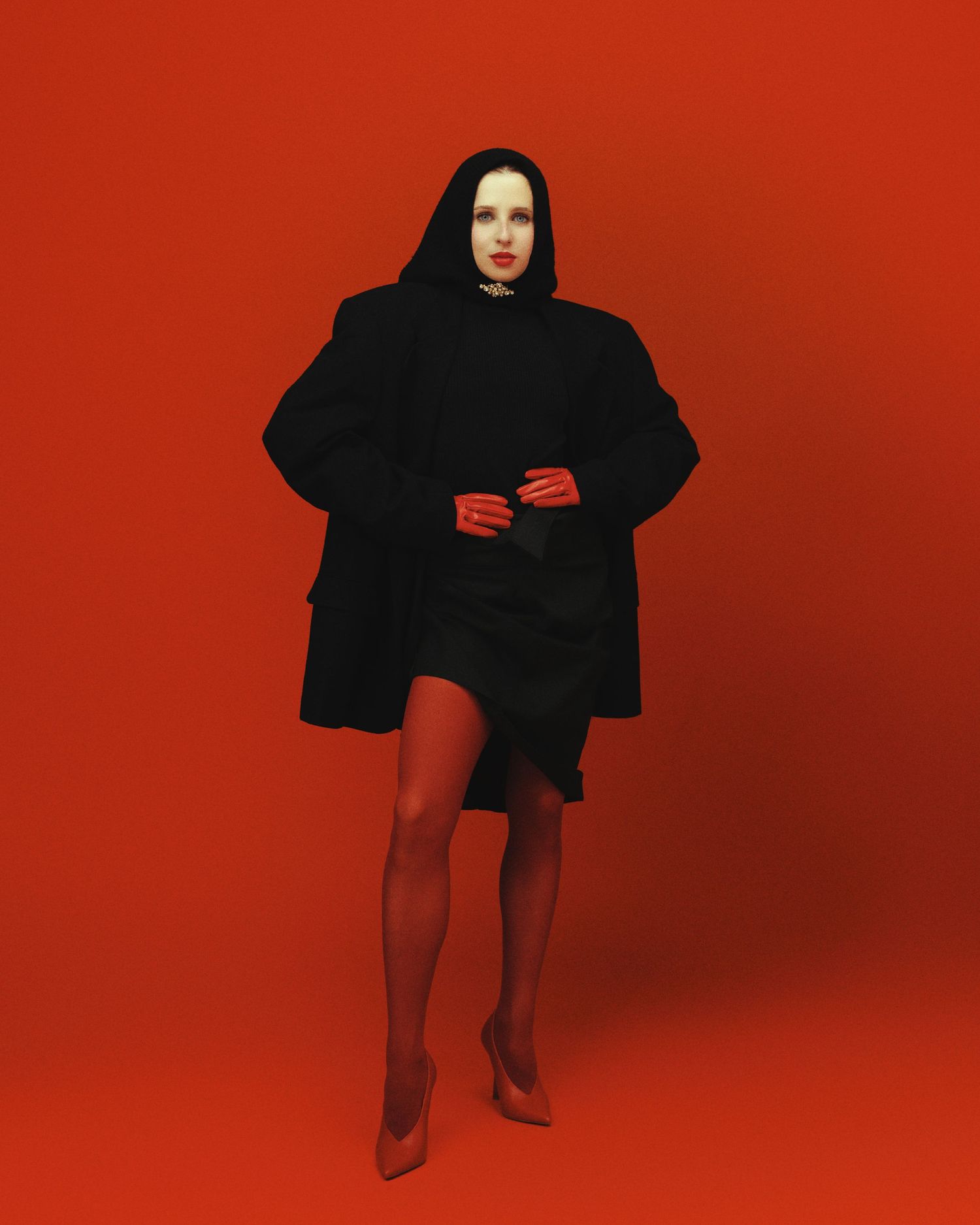 Woman in black coat, hood, and red gloves poses against bold red background wearing red heels.
