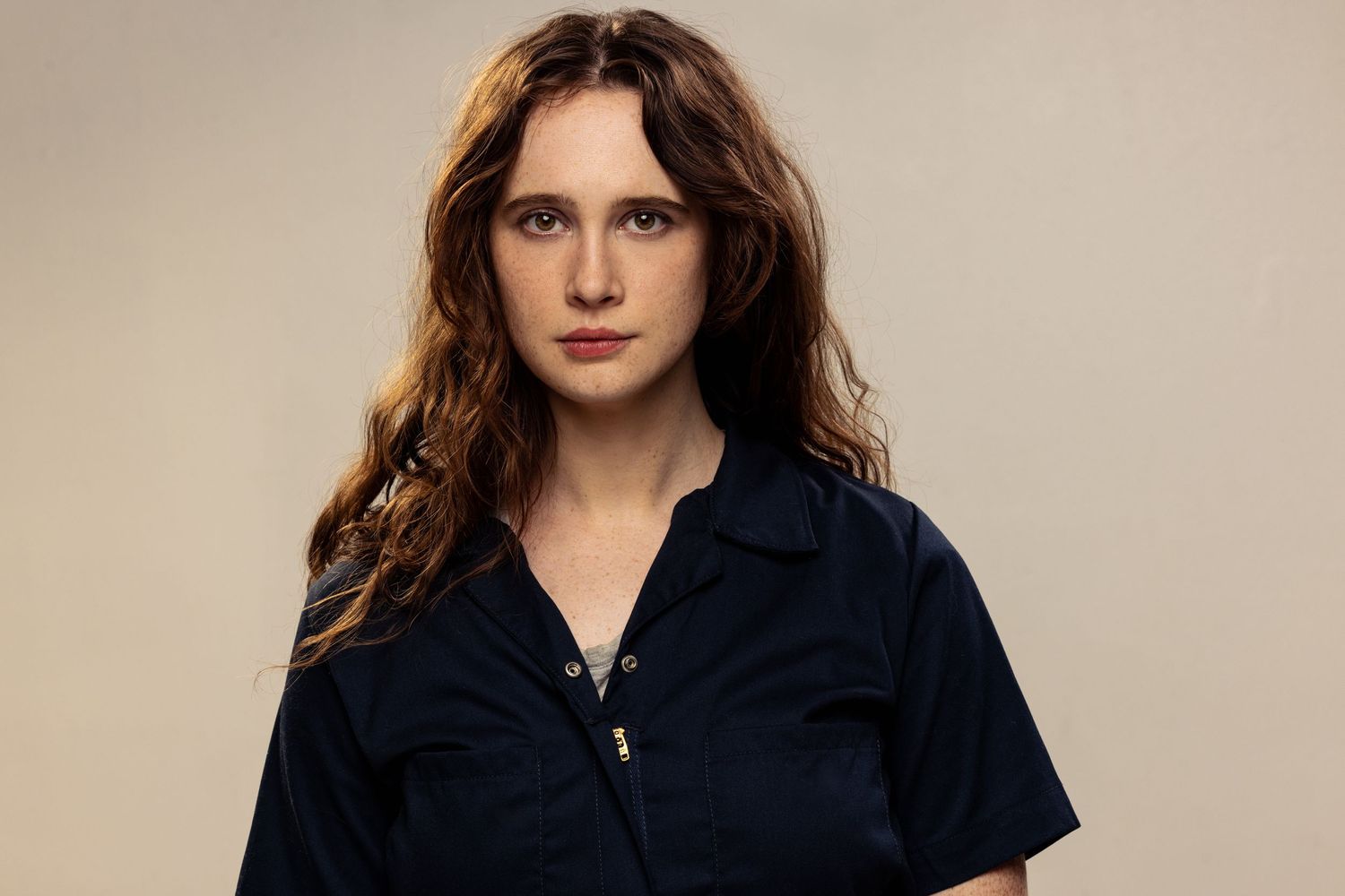 Young woman with wavy auburn hair wearing a navy blue jumpsuit, posing against a neutral beige background.