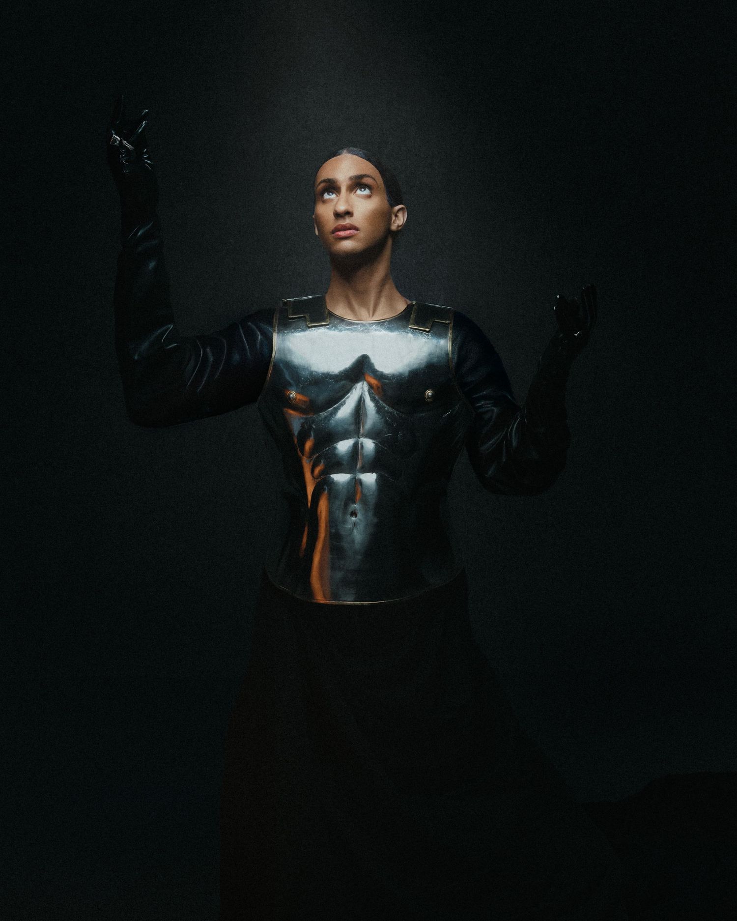 Person wearing a metallic superhero chest armor top with black gloves reaches upward in dark studio.