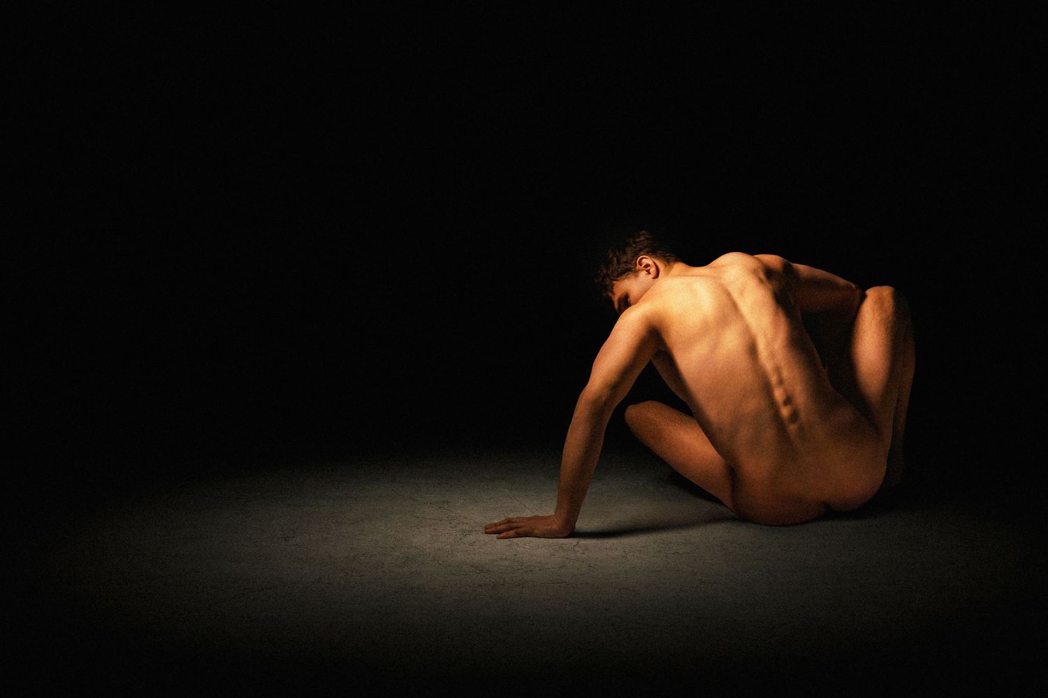 Nude male figure curled on floor in dramatic chiaroscuro lighting against black background.