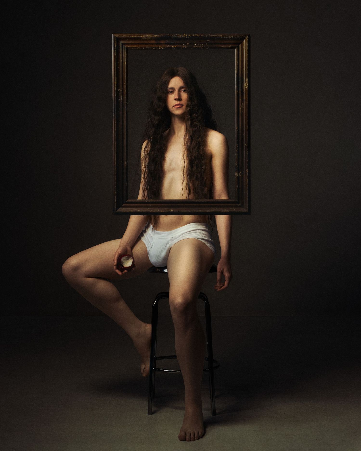Person sitting on stool holding ornate empty picture frame in front of face in studio portrait.