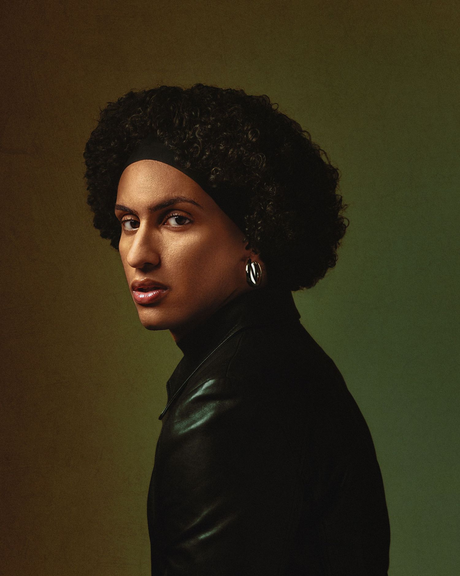 Close-up portrait of person with natural curly hair and hoop earring against green gradient background.