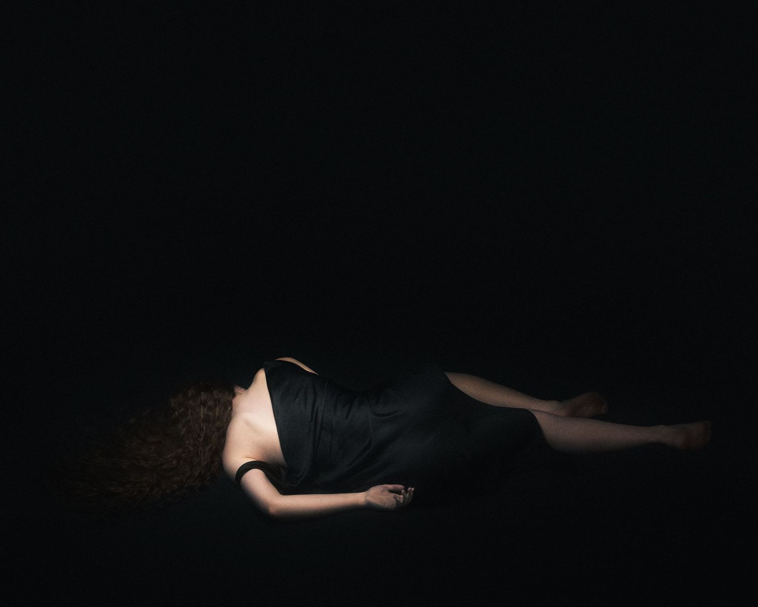 Woman in black dress lying on dark floor in dramatic low-key lighting photography.
