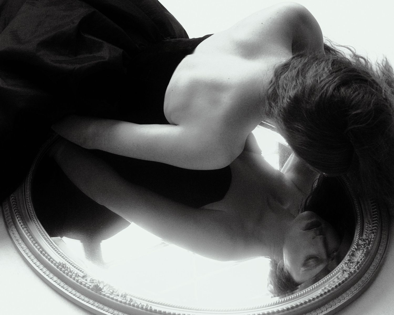 Black and white photo of a person lying over an ornate mirror reflecting their face.