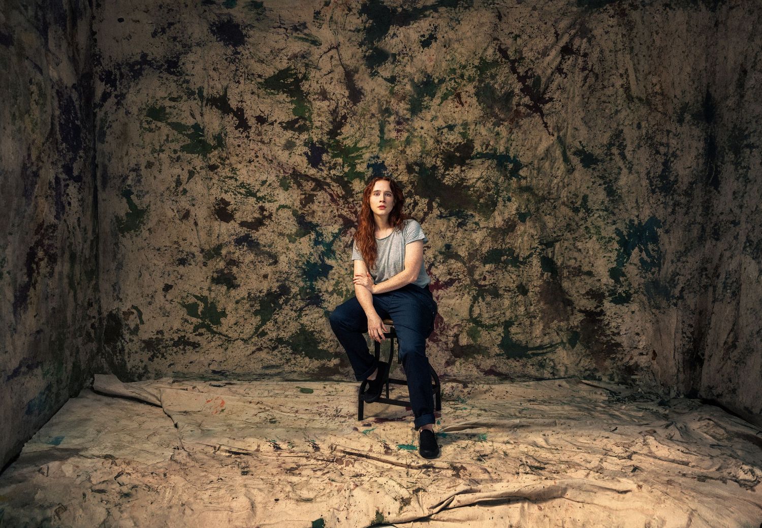 Woman sitting on chair in rustic, paint-splattered room with textured walls and debris-covered floor.