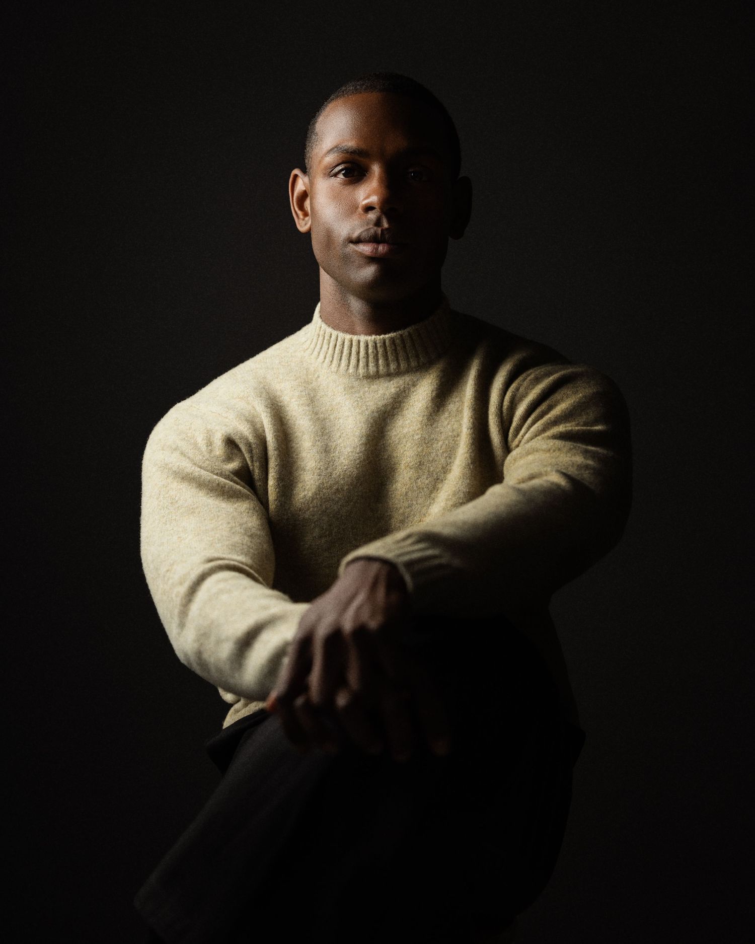 A man in a beige knit sweater sits confidently against a dark background in a dramatic studio portrait.