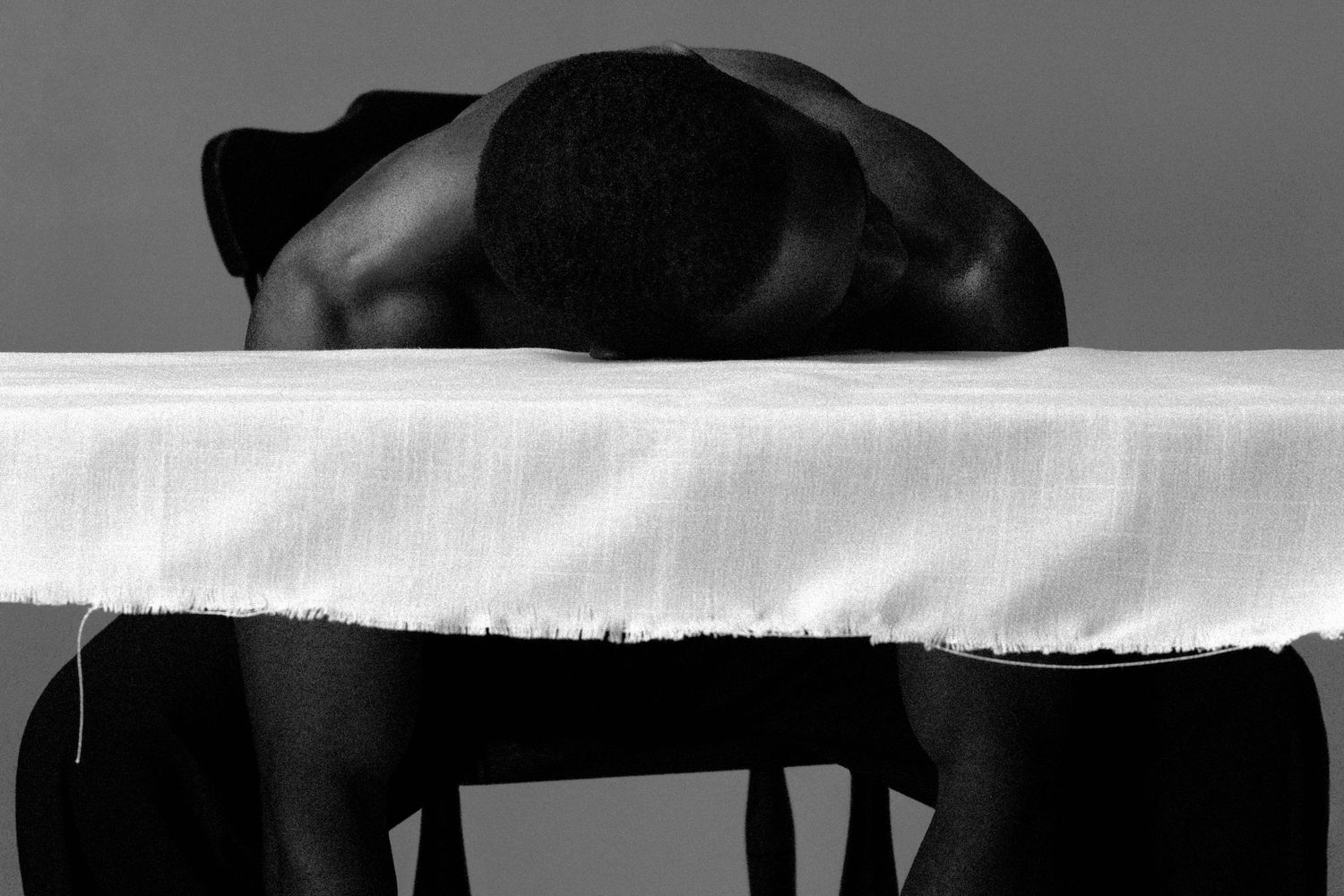 Black and white photo of shirtless person resting head on white cloth-covered table.