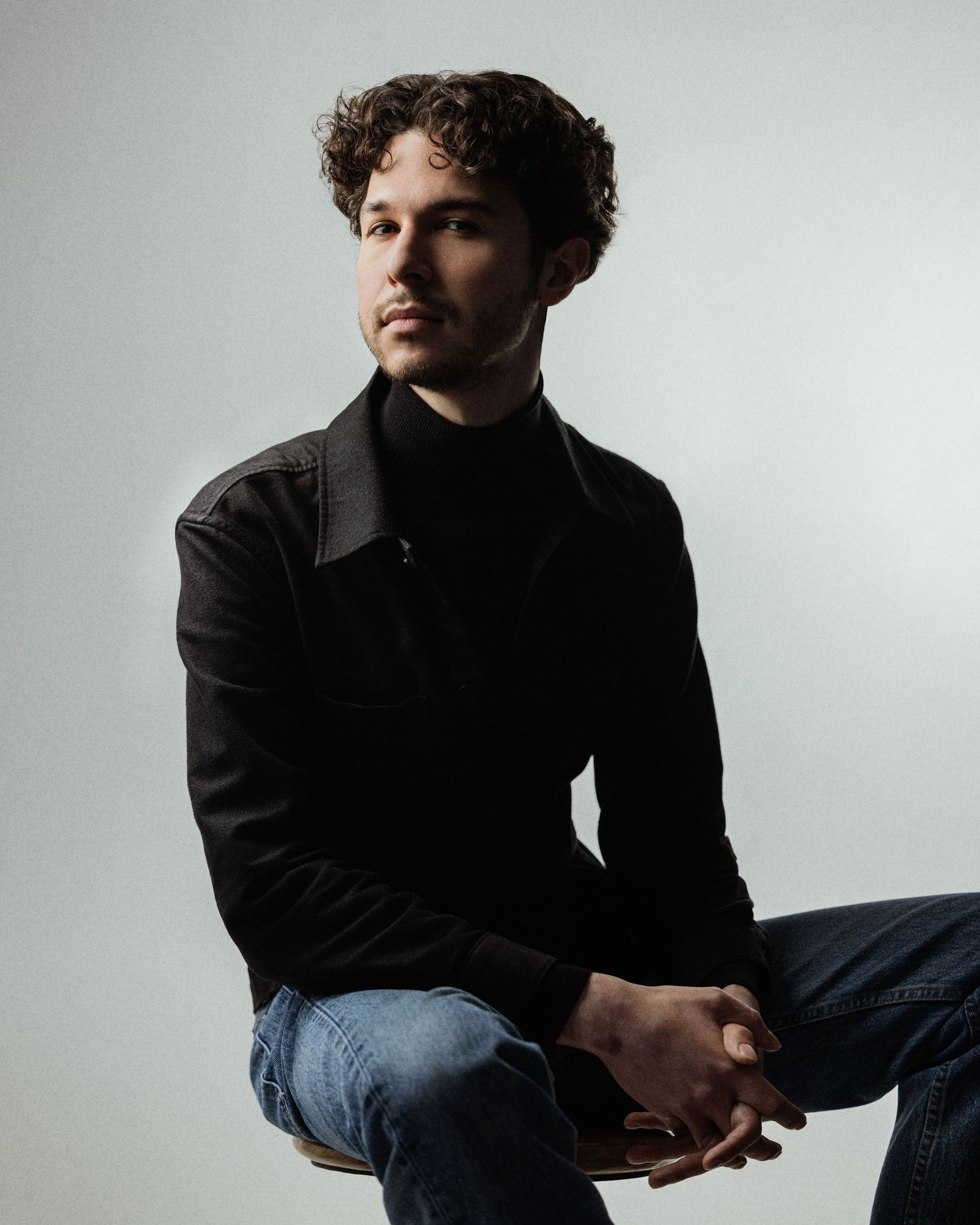 Young man with curly hair in black turtleneck jacket and jeans sitting thoughtfully against gray background.