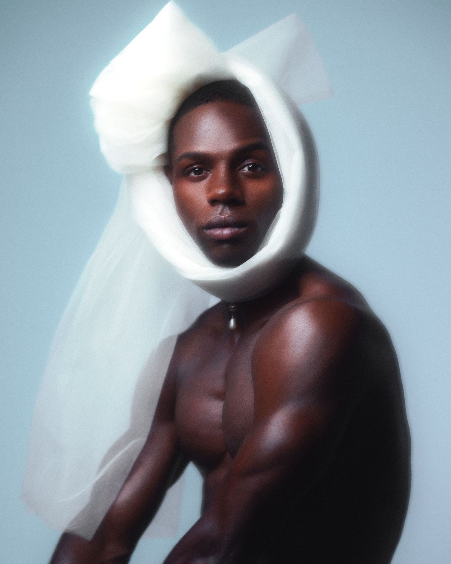 Shirtless Black man wearing white tulle veil headpiece in artistic studio portrait.