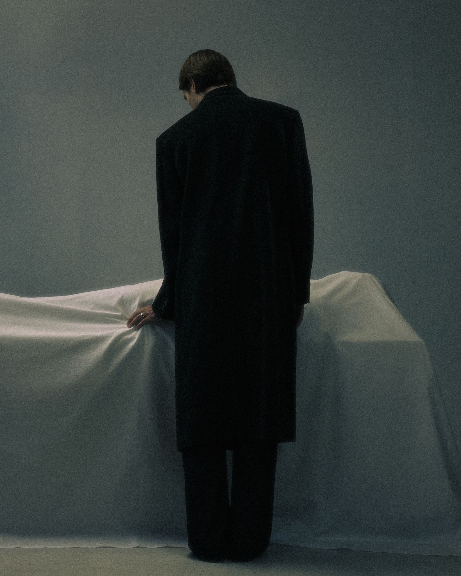 Person in a long black coat standing with their back turned toward a white draped table.