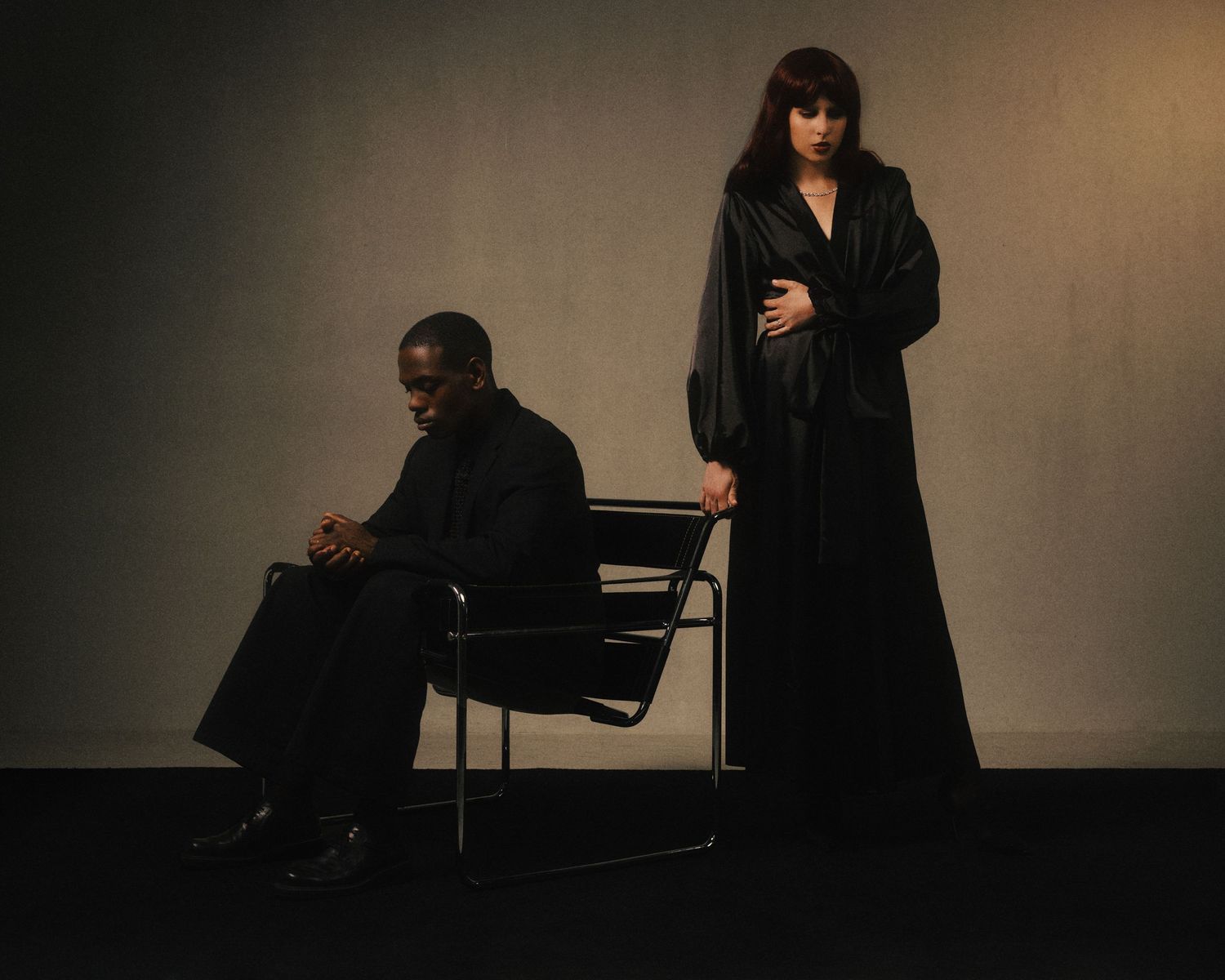 Two people dressed in black, one seated and one standing, posed against a gray studio background.