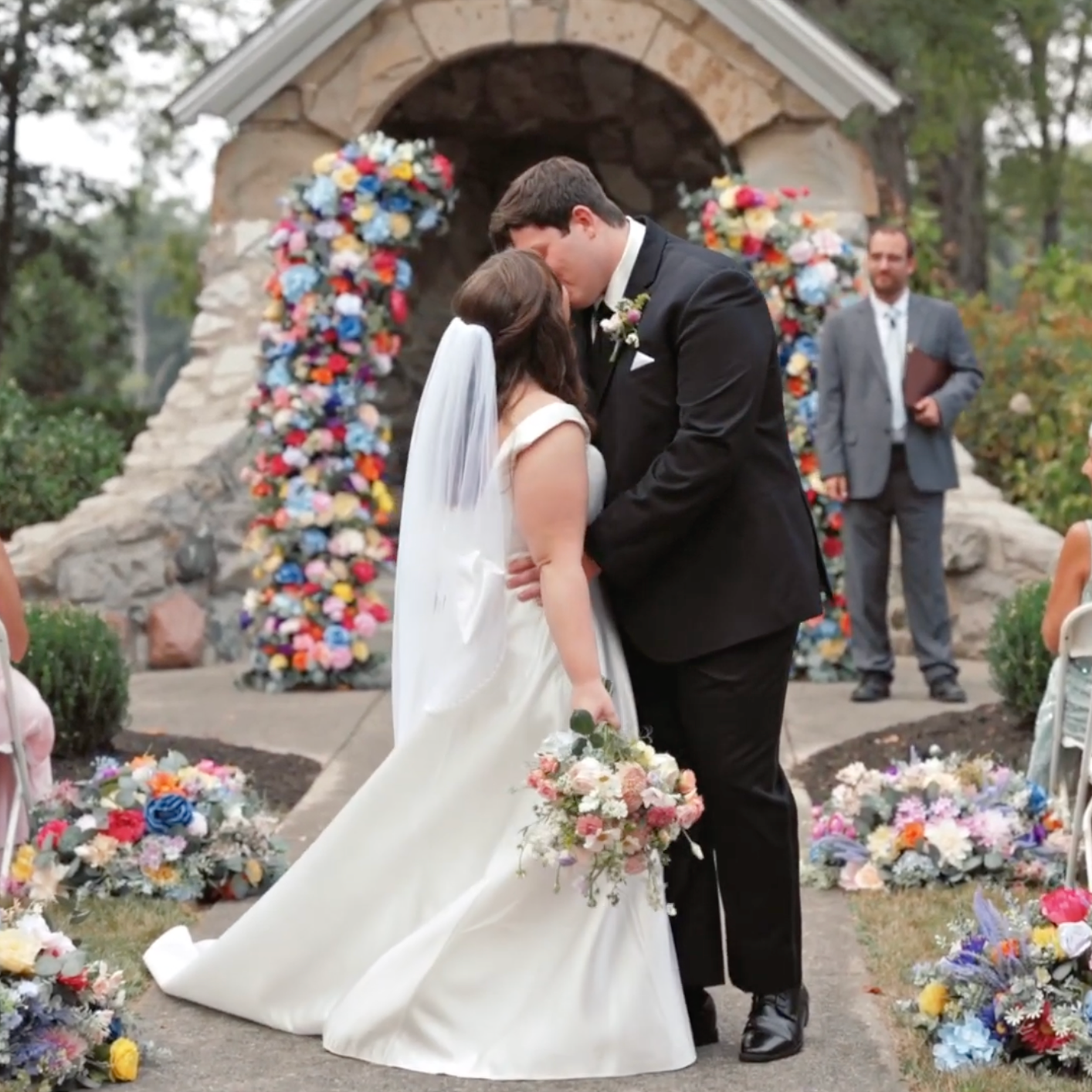 A couple shares a tender moment under a stone archway decorated with vibrant red and blue flowers at their wedding ceremony.