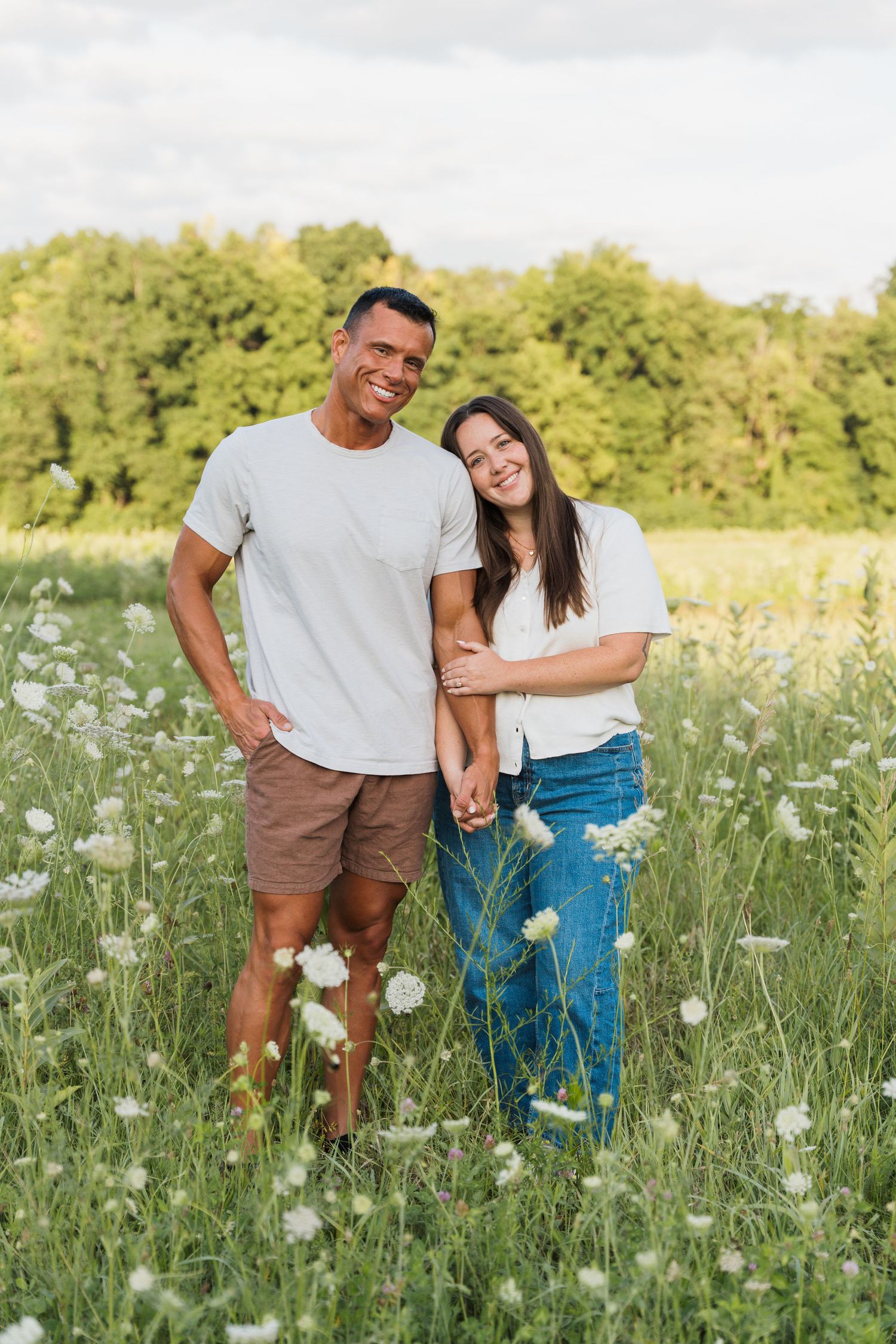 Happy couple holding hands in a wildflower meadow, wearing casual white shirts, with lush green trees in the background.