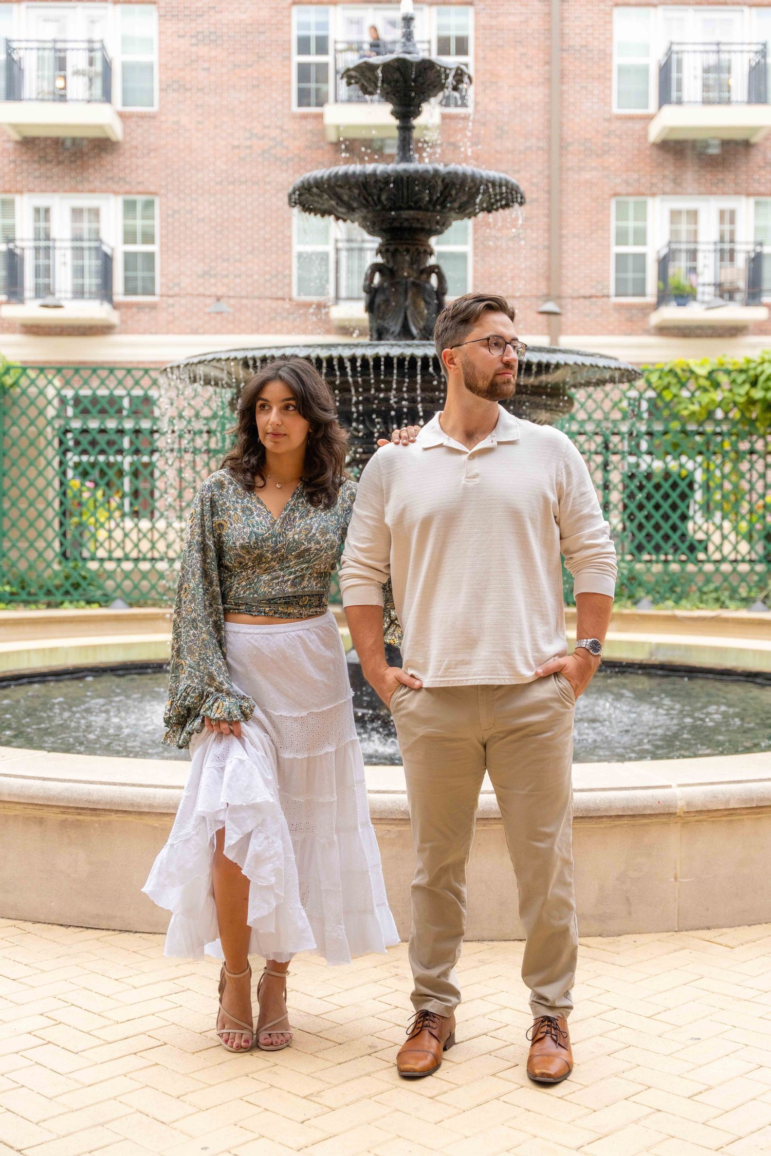 Couple in coordinating white and beige outfits pose together by an ornate stone fountain in a garden setting.