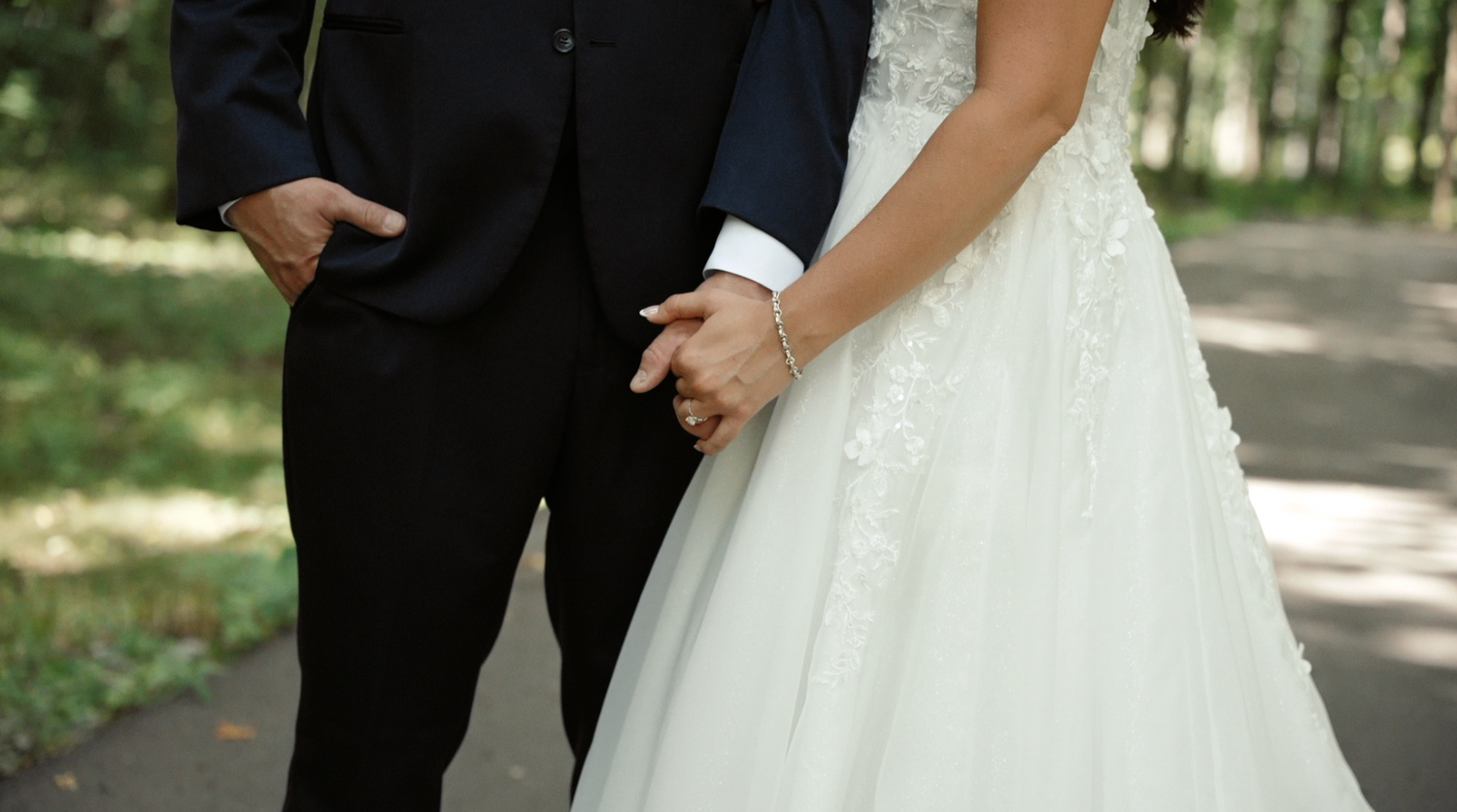 A close-up detail shot of a bride and groom's hands holding each other outdoors in a natural setting.