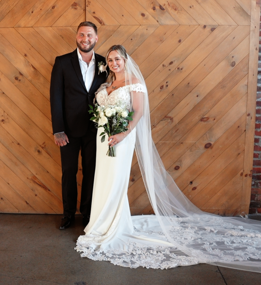 A couple in wedding attire poses against a wooden chevron wall backdrop with a long lace veil and white bouquet.
