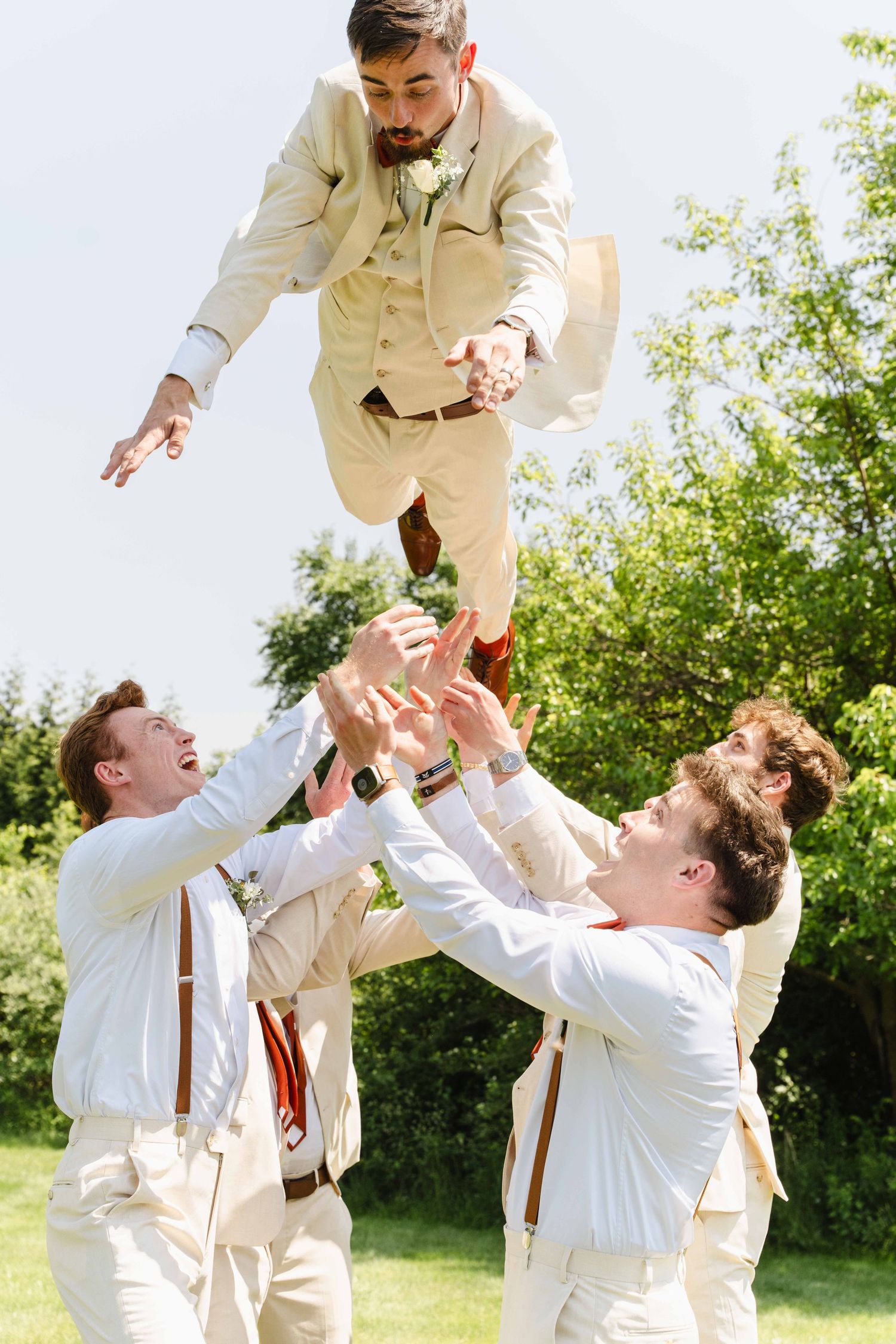 Groomsmen in cream suits tossing groom into the air during outdoor wedding celebration on sunny day.