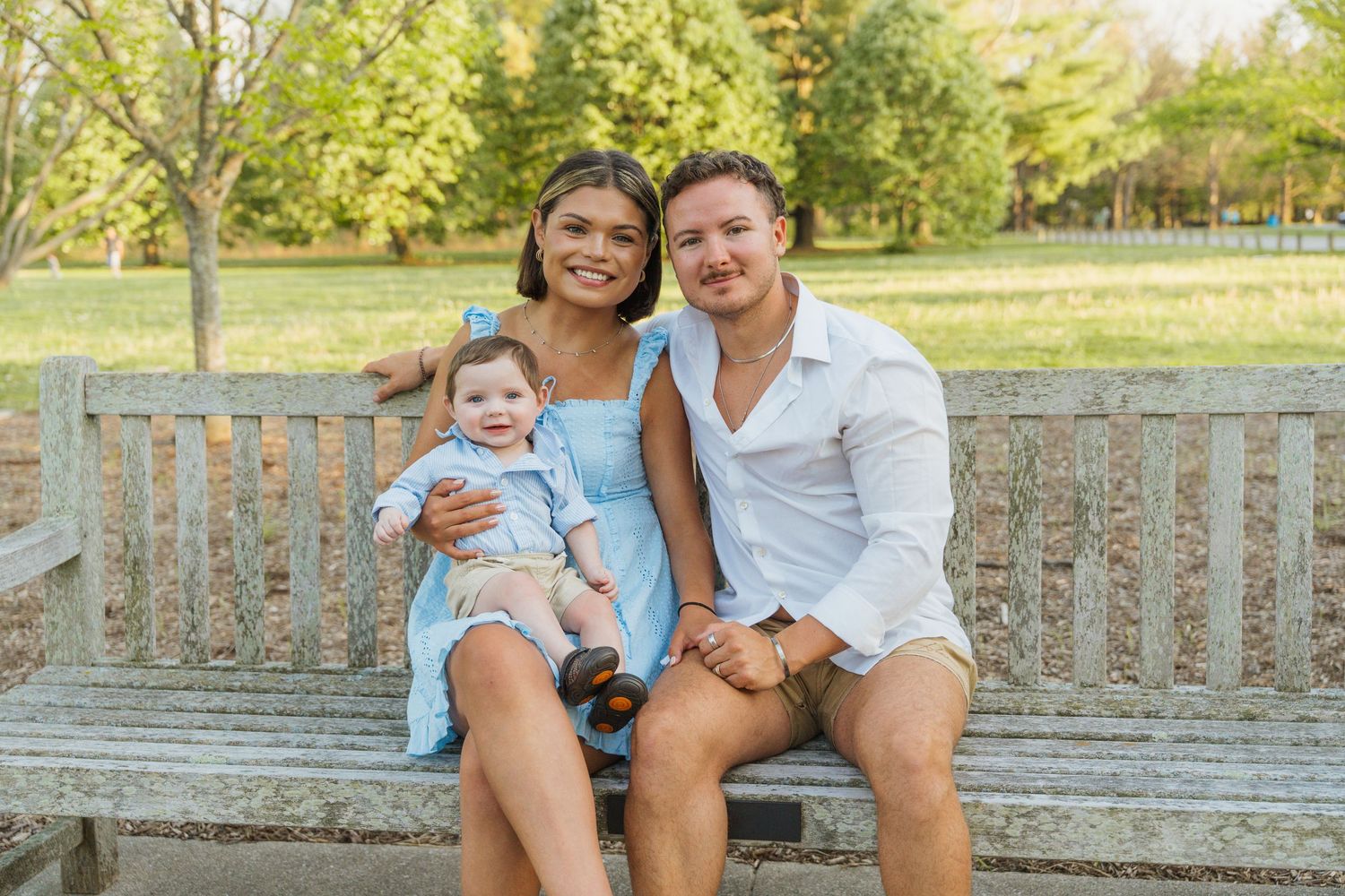 Family of three smiling on a wooden park bench, mother holding baby in blue outfit, father beside them, green trees background.