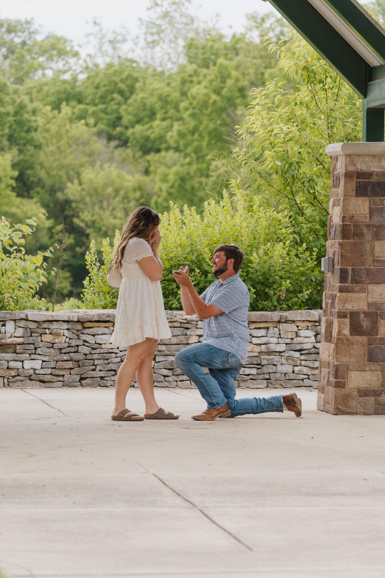 Man kneeling on one knee proposing to woman in white dress outdoors near stone wall and lush greenery.