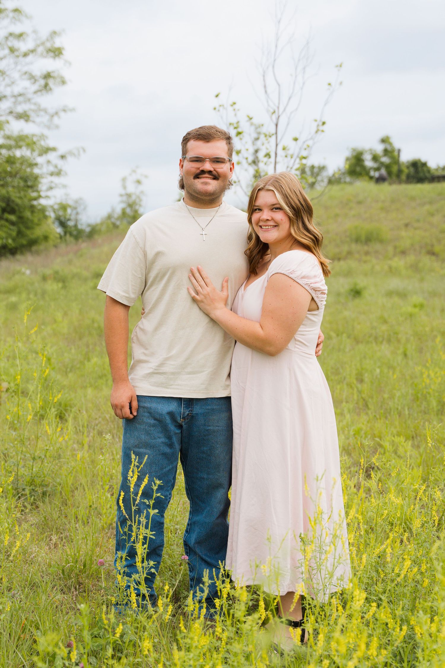 A smiling couple poses together in a green meadow, the man in jeans and the woman in a pink dress.
