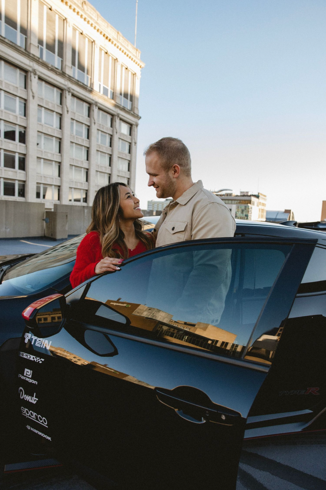 A couple shares an intimate moment while leaning against a black car outside a modern building.