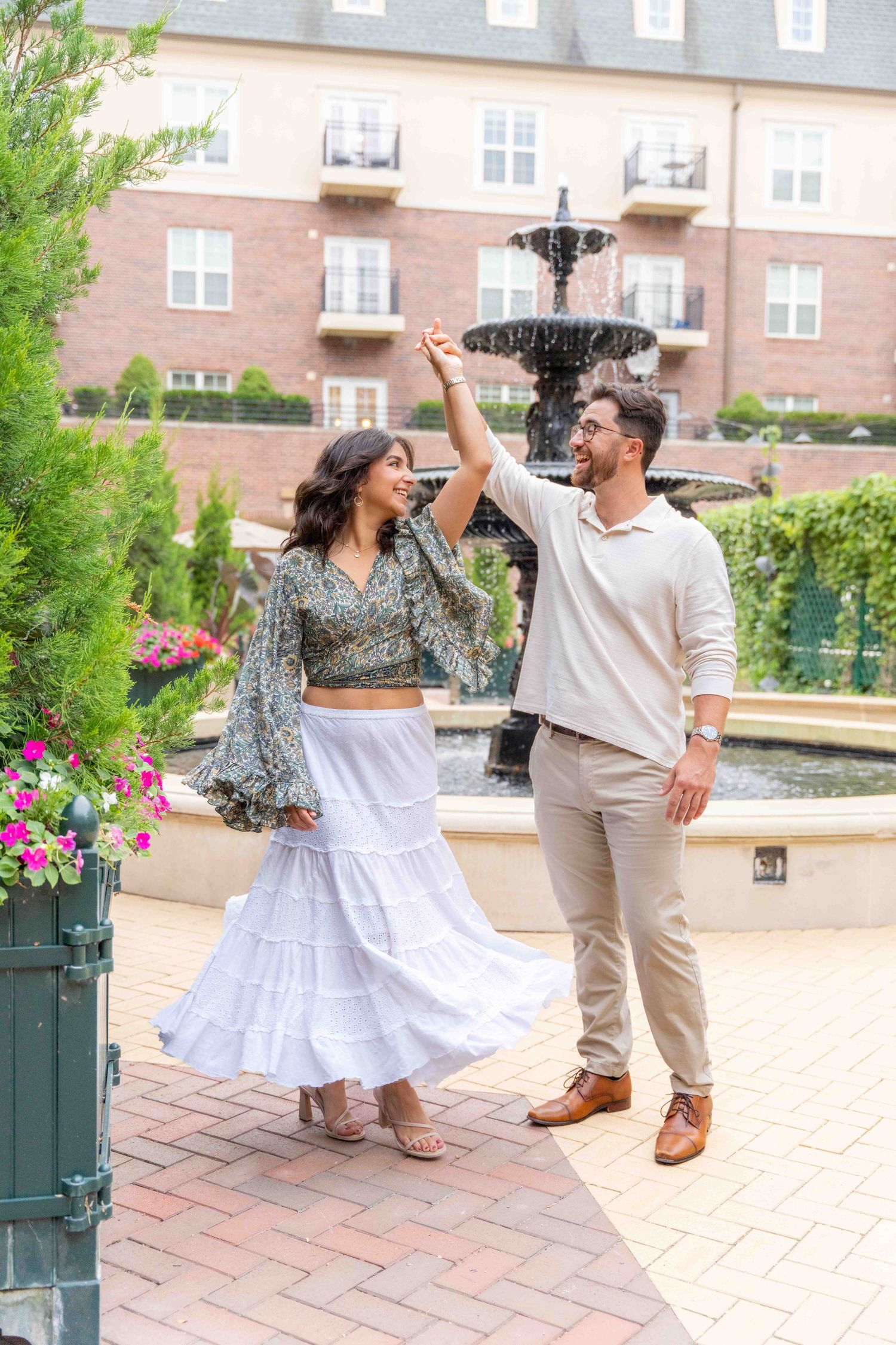 A couple dances joyfully together on a brick patio surrounded by flowers and architecture.