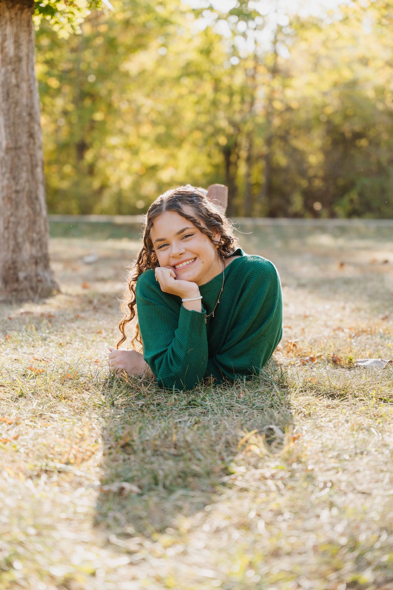 Smiling young woman with long curly hair in a green sweater lying on a grassy lawn, resting her chin on her hand in warm autumn sunlight.