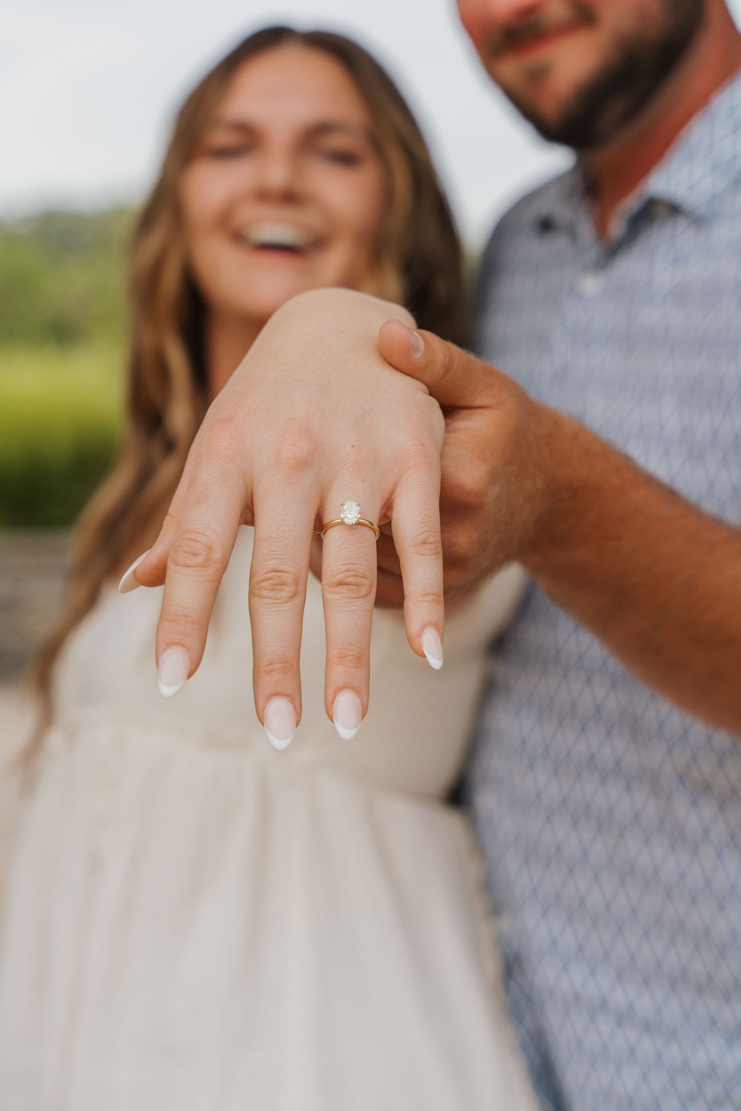 Smiling couple showing off engagement ring with oval diamond on woman's hand with white manicured nails.
