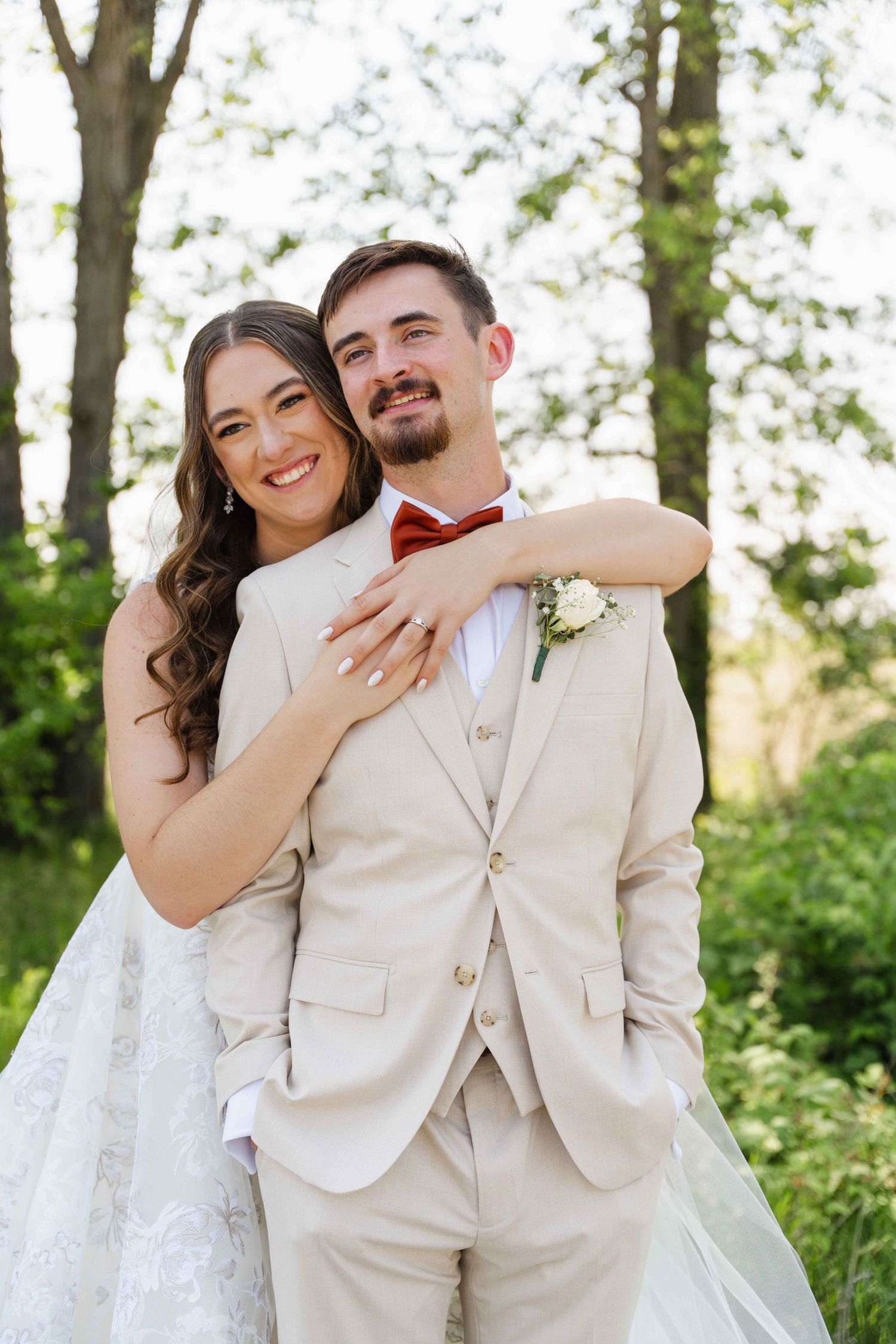 A smiling bride in a white lace gown embraces a groom in a beige suit with red bow tie outdoors.