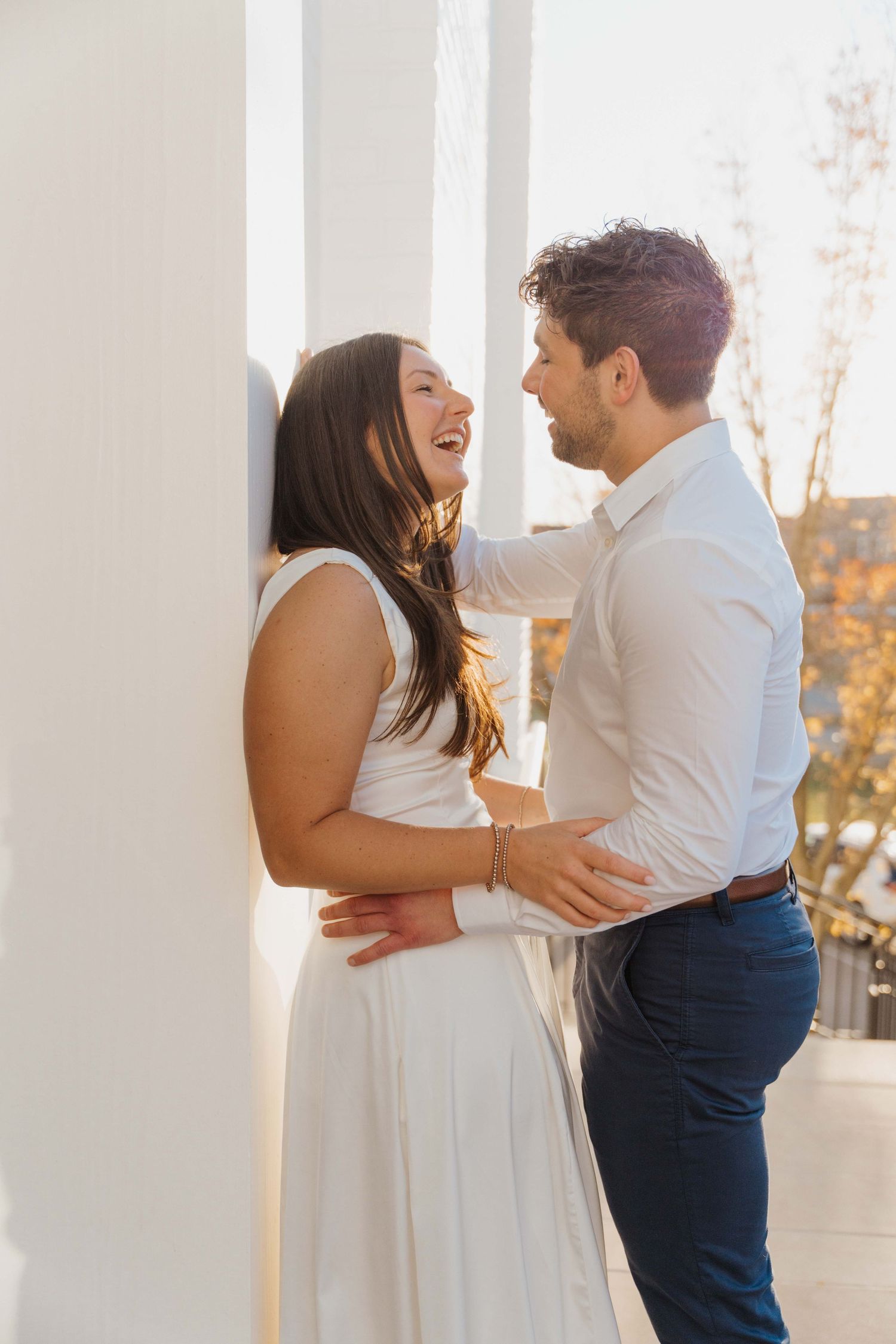 A couple shares a joyful moment together on a sun-lit porch, with the woman in white dress laughing while embracing.