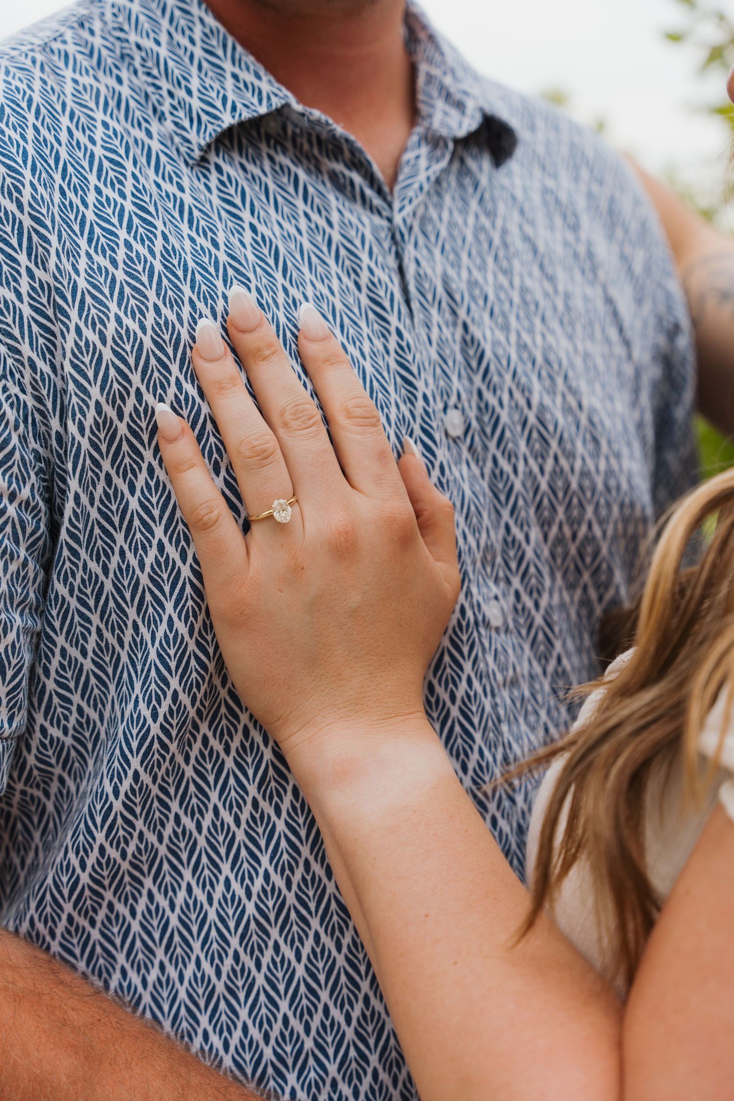 Woman displaying engagement ring with oval diamond on hand resting against man's blue patterned shirt.