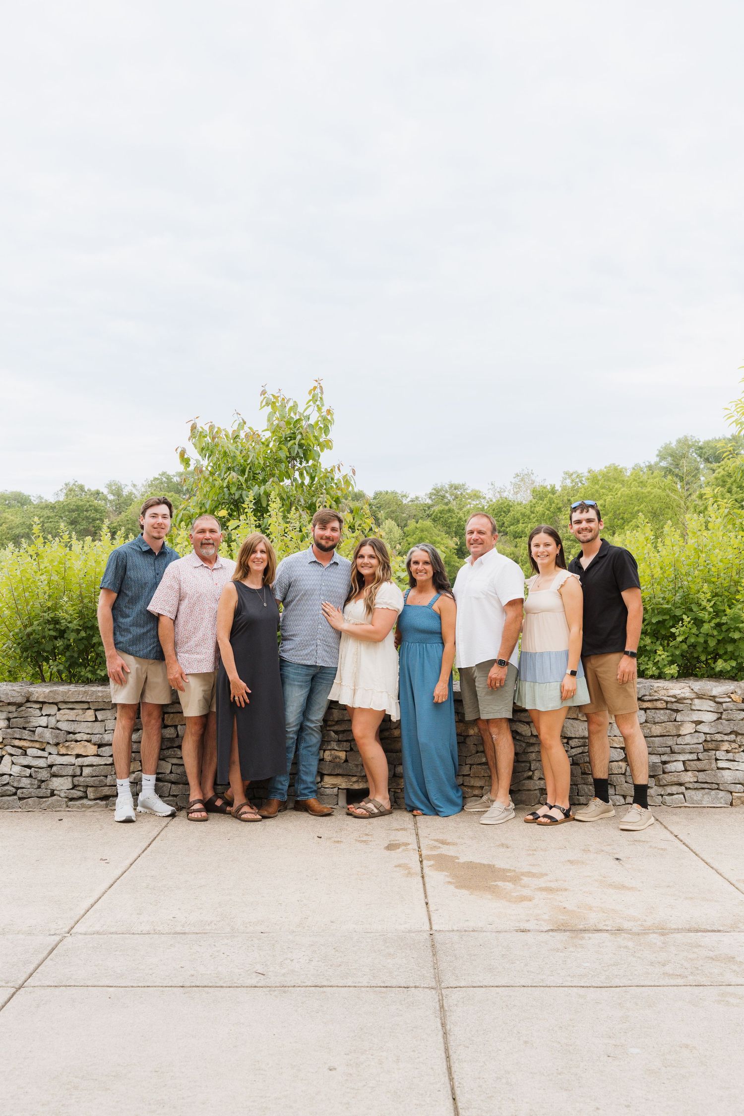 A group of eight adults posing together outdoors near a stone wall with lush green foliage in the background.