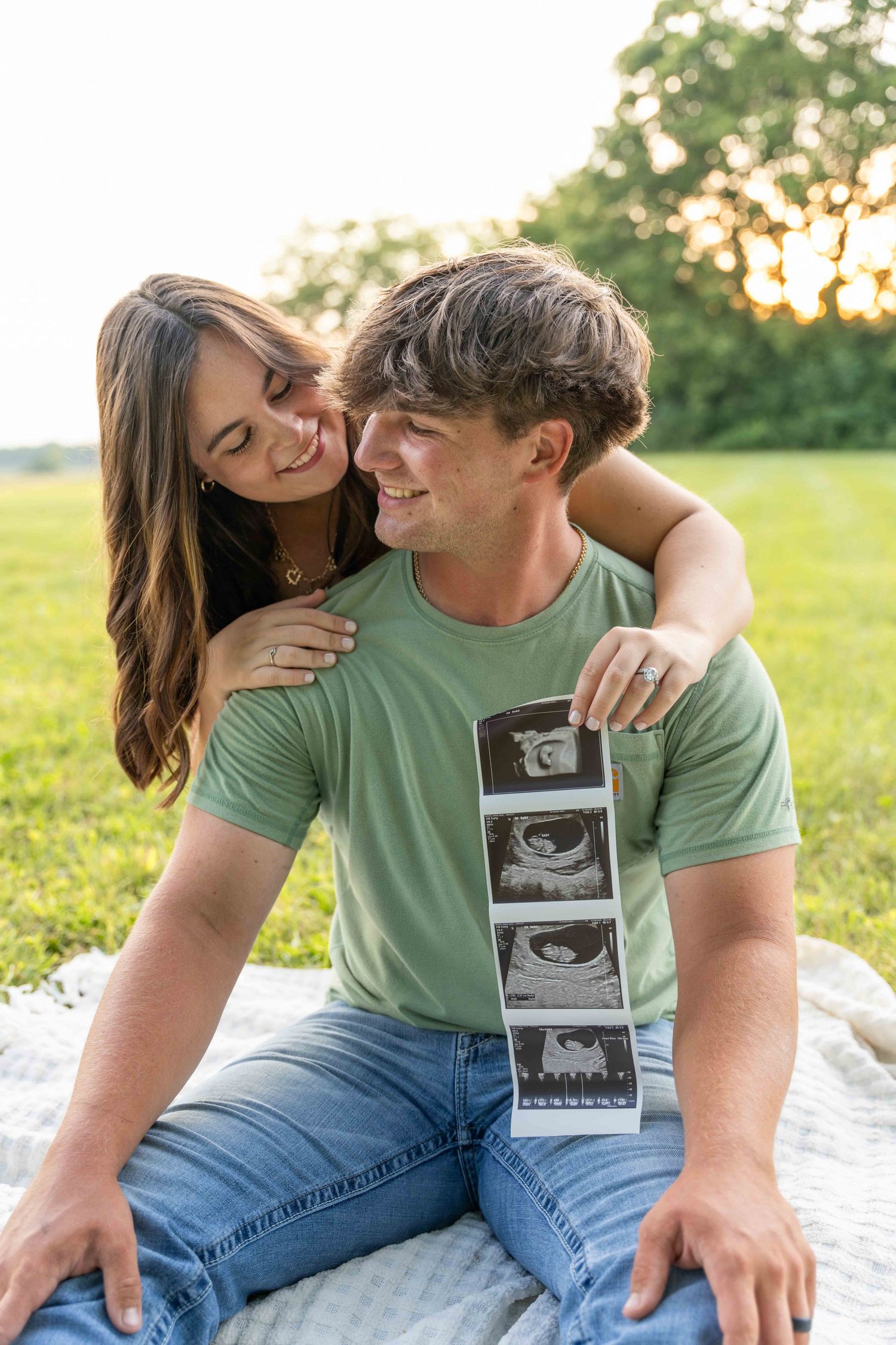 A couple sits on a blanket outdoors sharing ultrasound photos during a pregnancy announcement photoshoot at sunset.