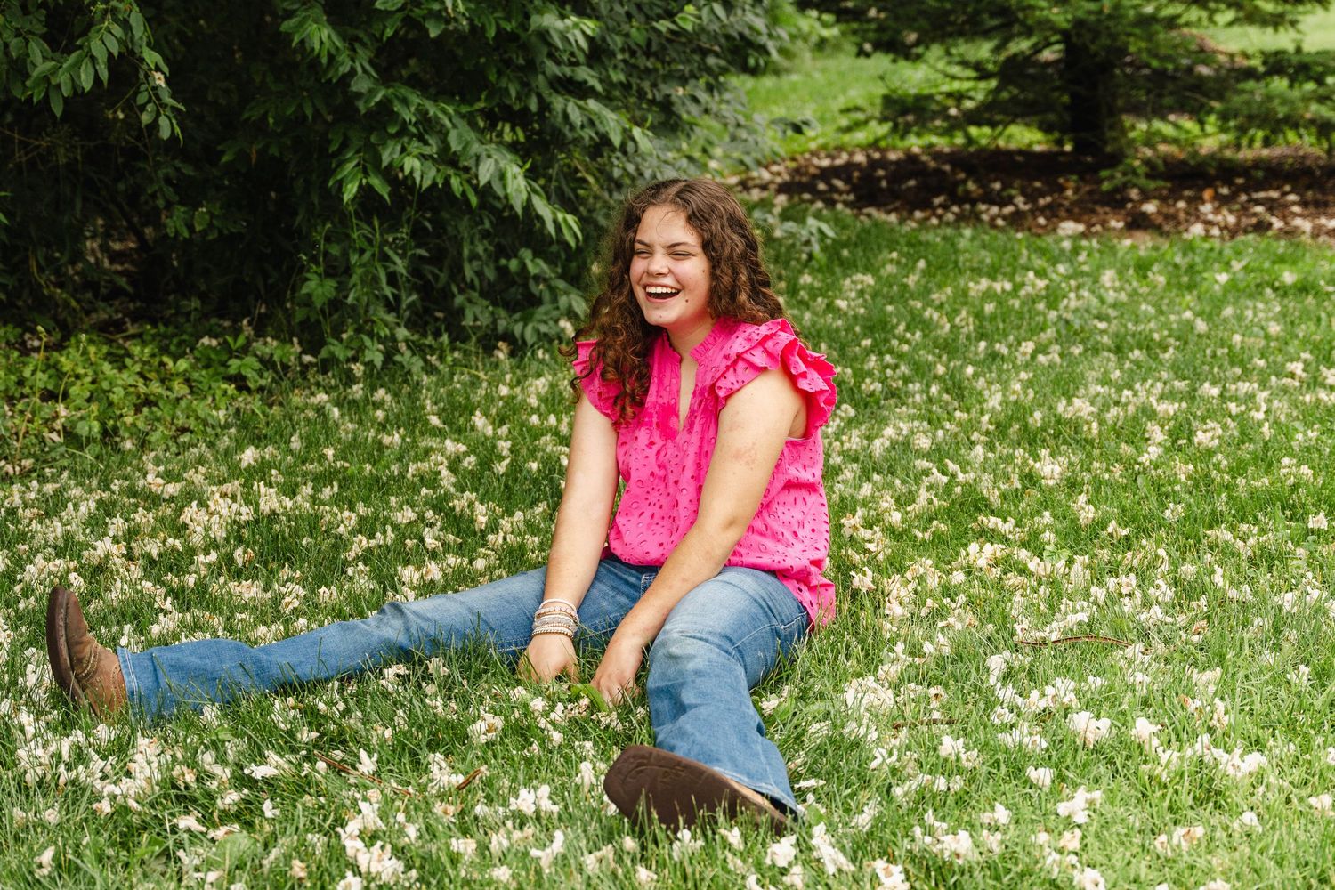 A person in a pink shirt and jeans sits joyfully among blooming white daisies in a lush green field.