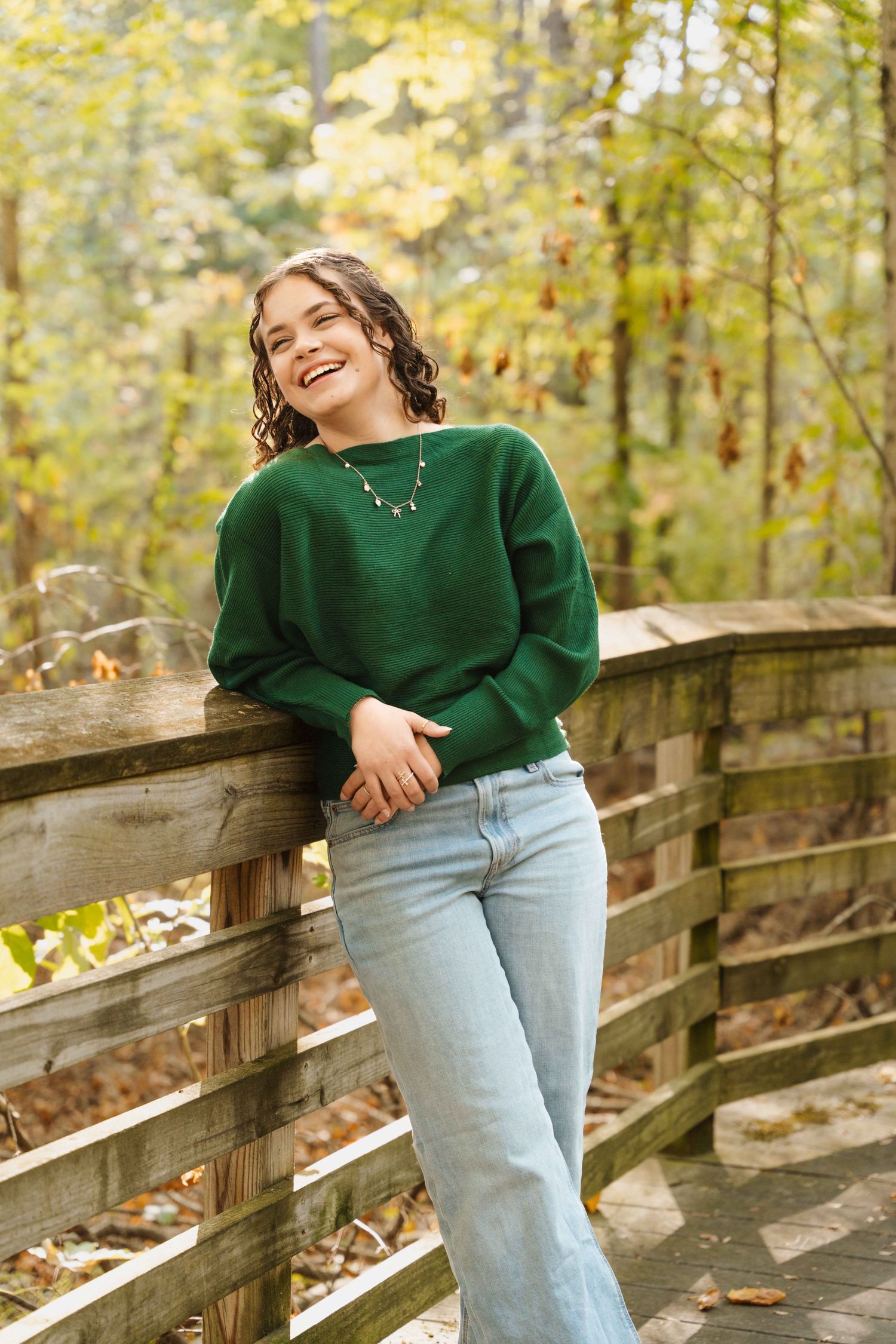 A person in a green sweater and light jeans leans against a wooden bridge railing surrounded by autumn foliage.