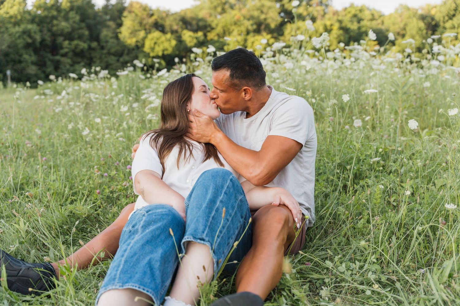 Couple sitting in a wildflower meadow sharing a romantic kiss wearing white shirts and jeans on a sunny day.
