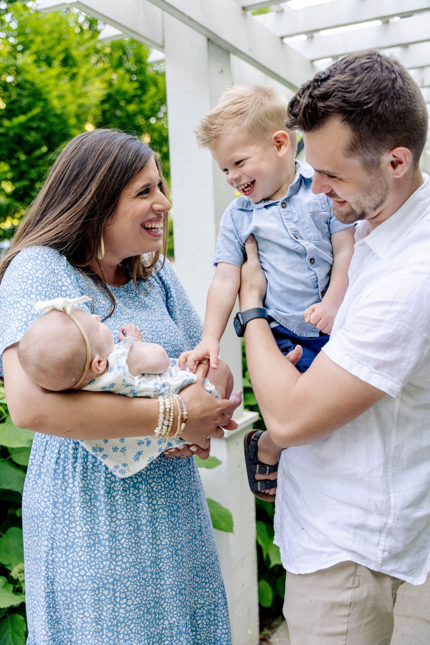 Happy family of four with parents holding a laughing toddler and newborn baby outdoors near a white pergola.