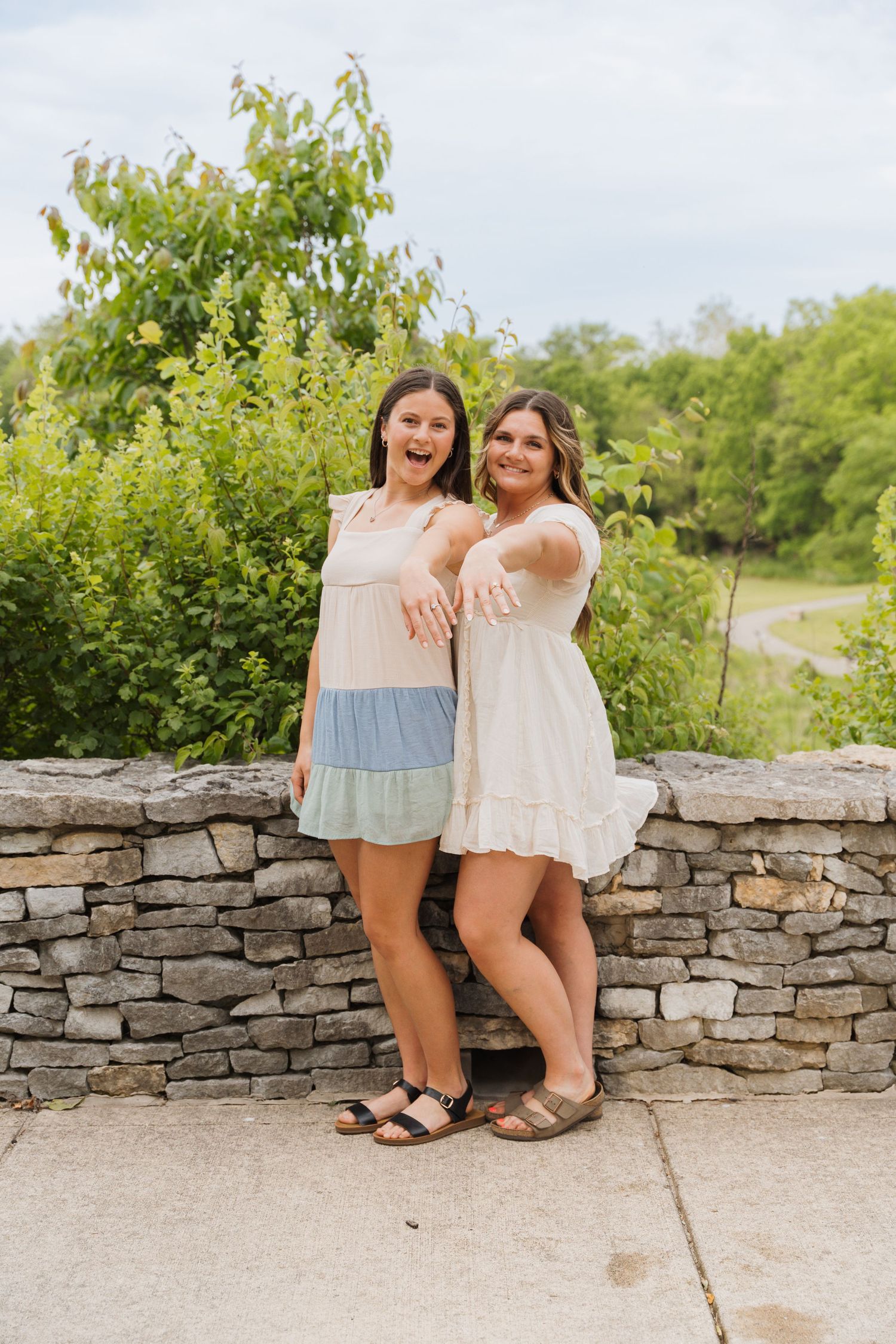 Two young women in summer dresses smiling and posing together near a stone wall with lush green trees behind them.