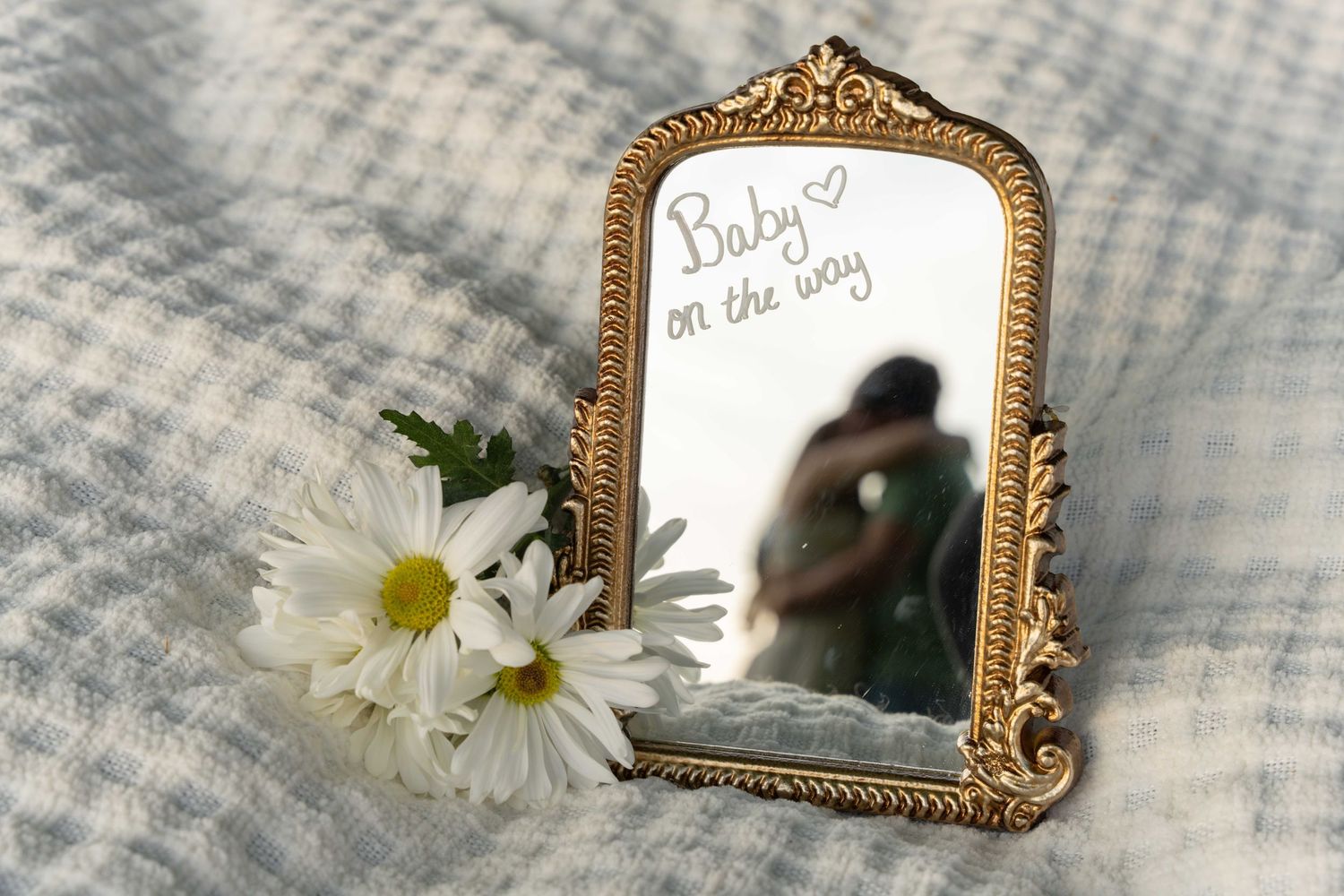 A vintage gold ornate mirror with white daisies and a handwritten note reflects a silhouetted figure on a soft gray blanket.