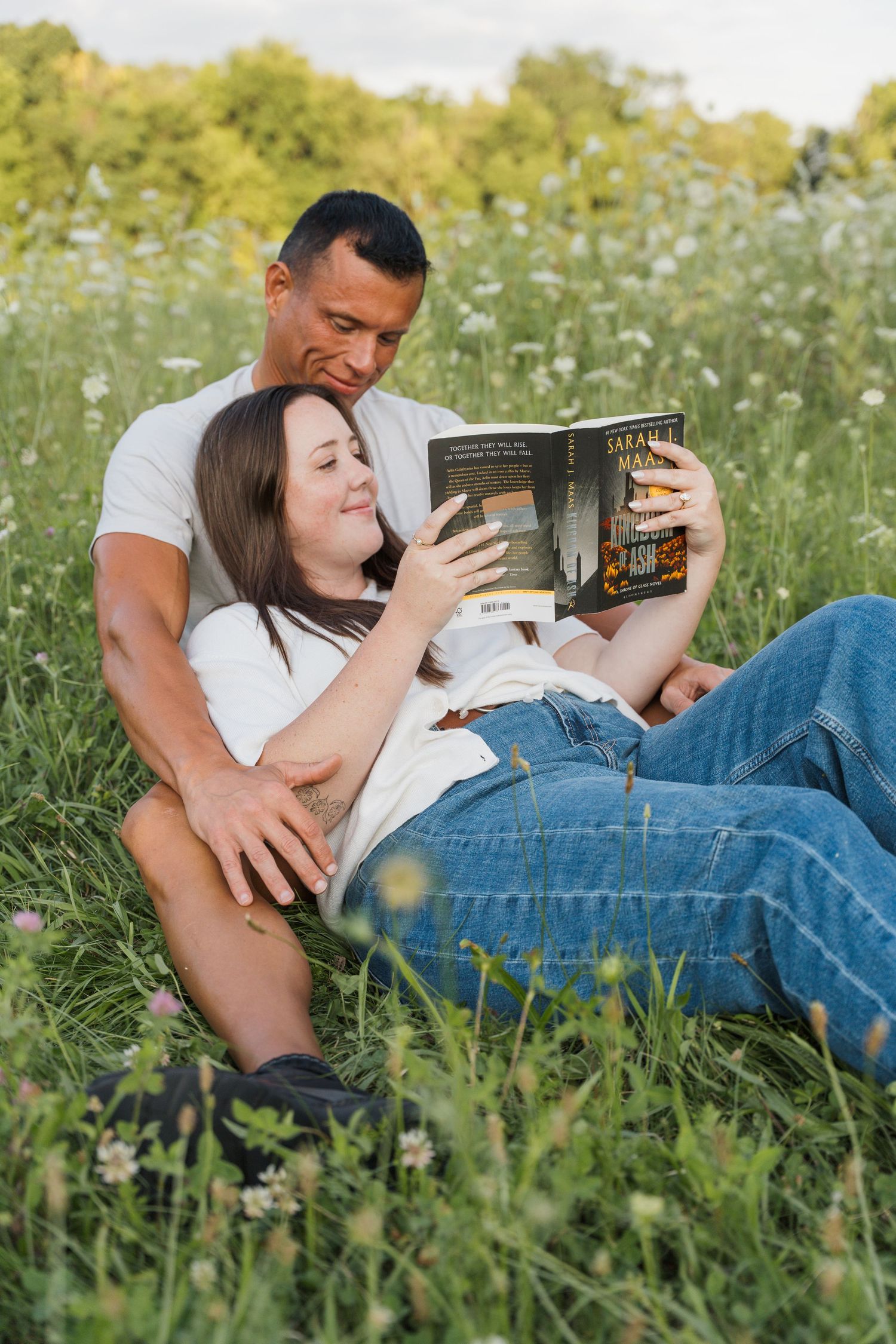 Couple relaxing in a wildflower meadow, woman reading a book while leaning against man in white tee and jeans.