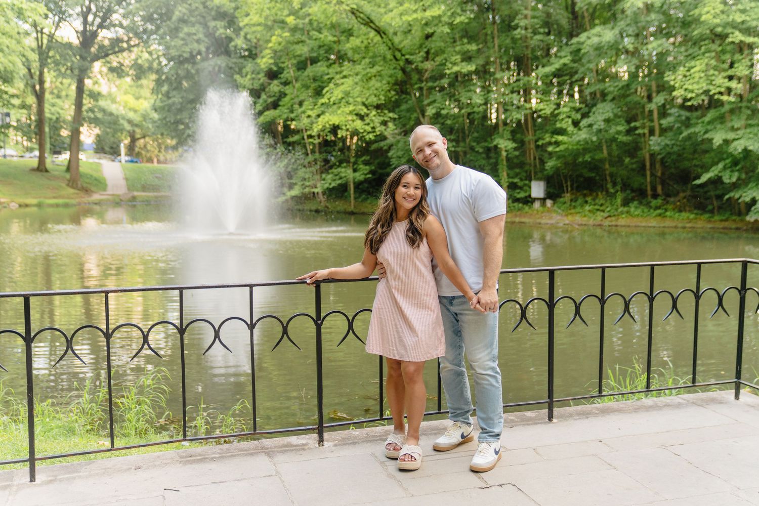 Couple smiling and holding hands by a decorative fence with a pond fountain and lush green trees in the background.