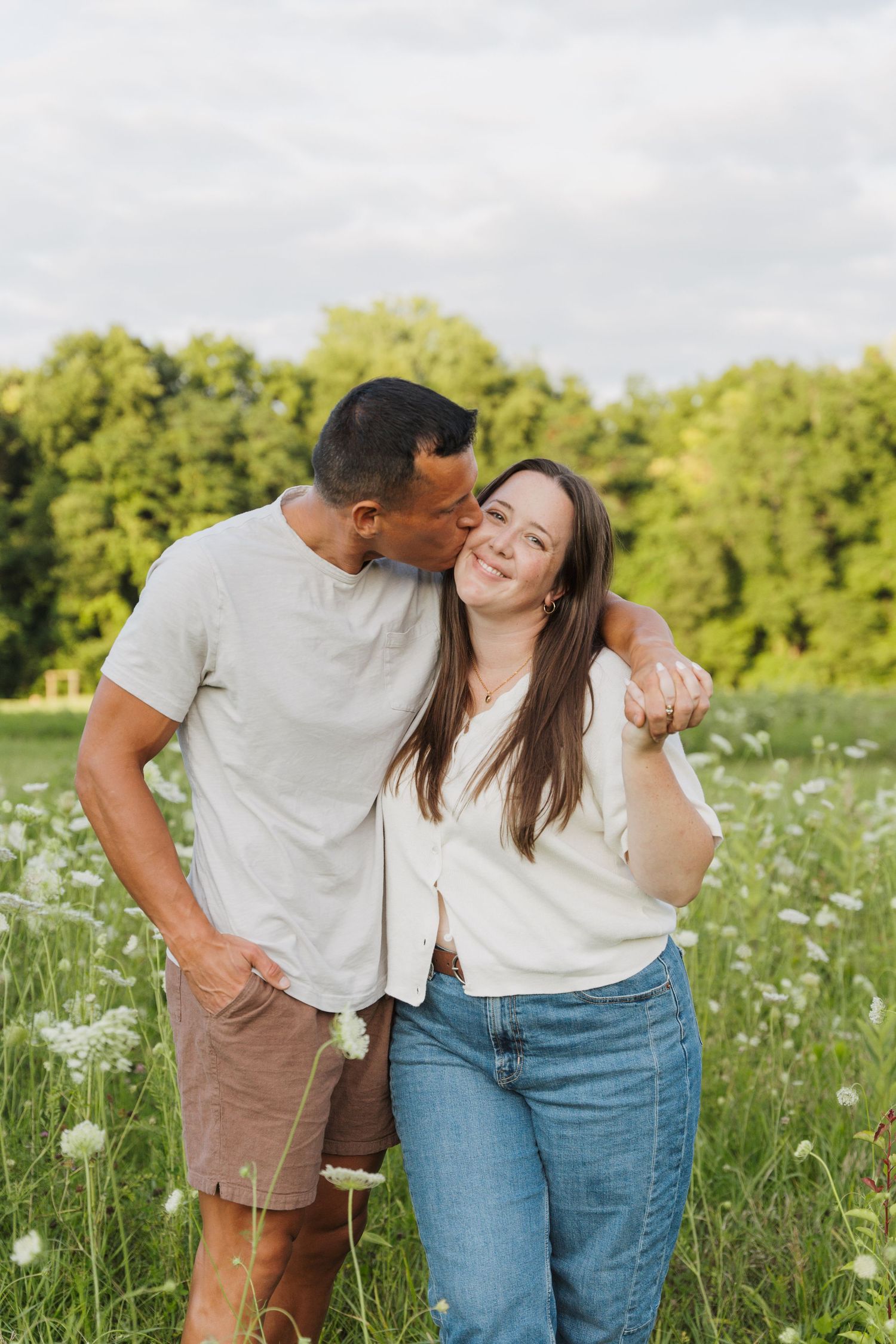 Couple sharing a romantic kiss in a wildflower meadow, man in beige shirt and shorts embracing smiling woman in jeans.
