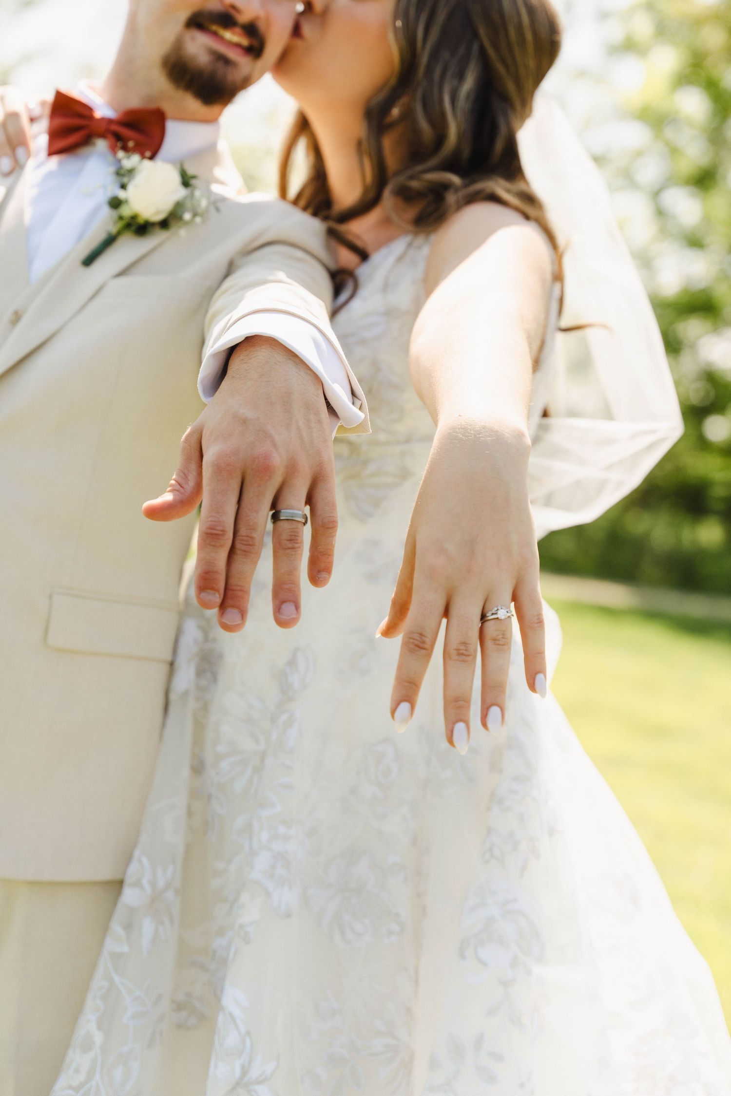 Close-up details of wedding rings and hands against a white lace dress in natural outdoor lighting.