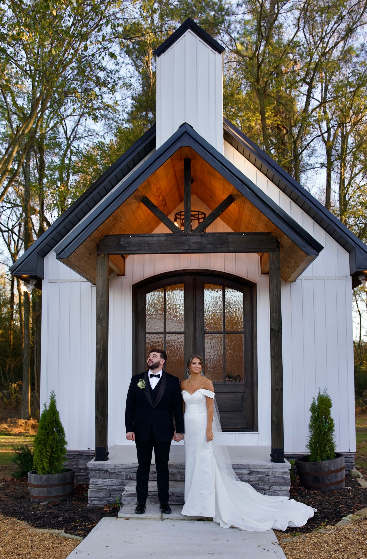 A couple poses in front of a charming white chapel with wooden accents and stone foundation in a wooded setting.